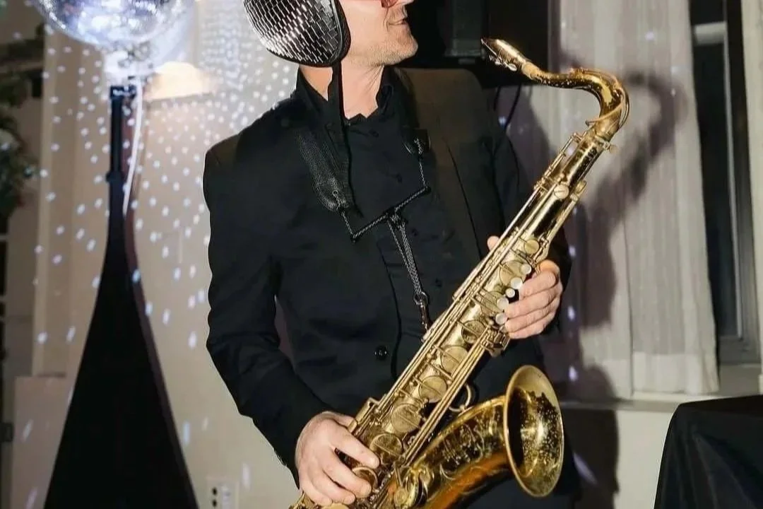 A man dressed in black holding a gold saxophone, with a black microphone near his face, in a warmly lit room with curtains and decorative lights.