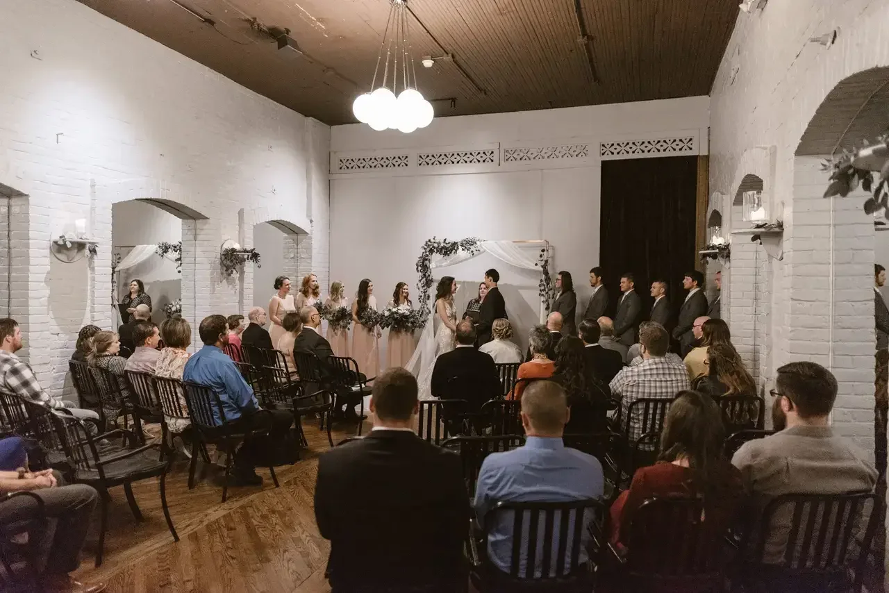 Indoor wedding ceremony with a bride and groom exchanging vows in front of an officiant, surrounded by bridesmaids and groomsmen, with guests seated in chairs facing the couple.