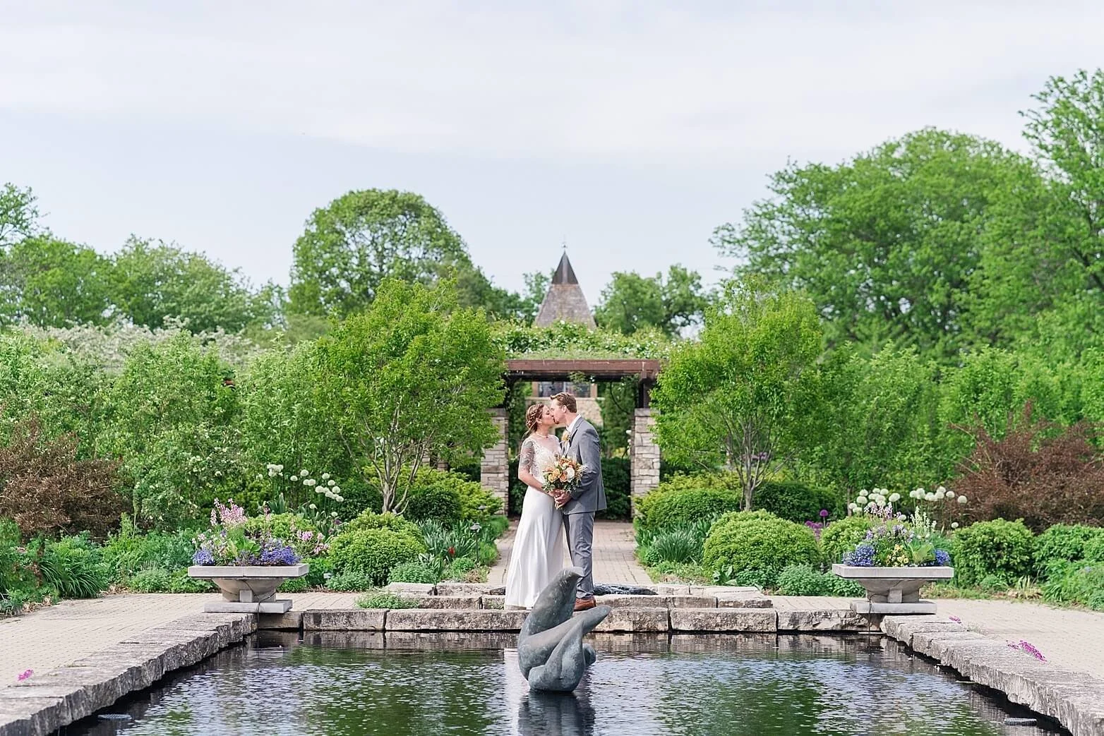 A bride and groom kissing in a garden with lush greenery and flowering plants, standing at the edge of a pond with a dolphin-shaped sculpture in the center.