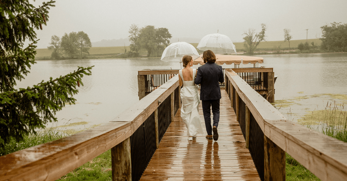 Couple walking on wooden dock towards a boat, holding umbrellas in rainy weather, overlooking a lake with trees in the background.