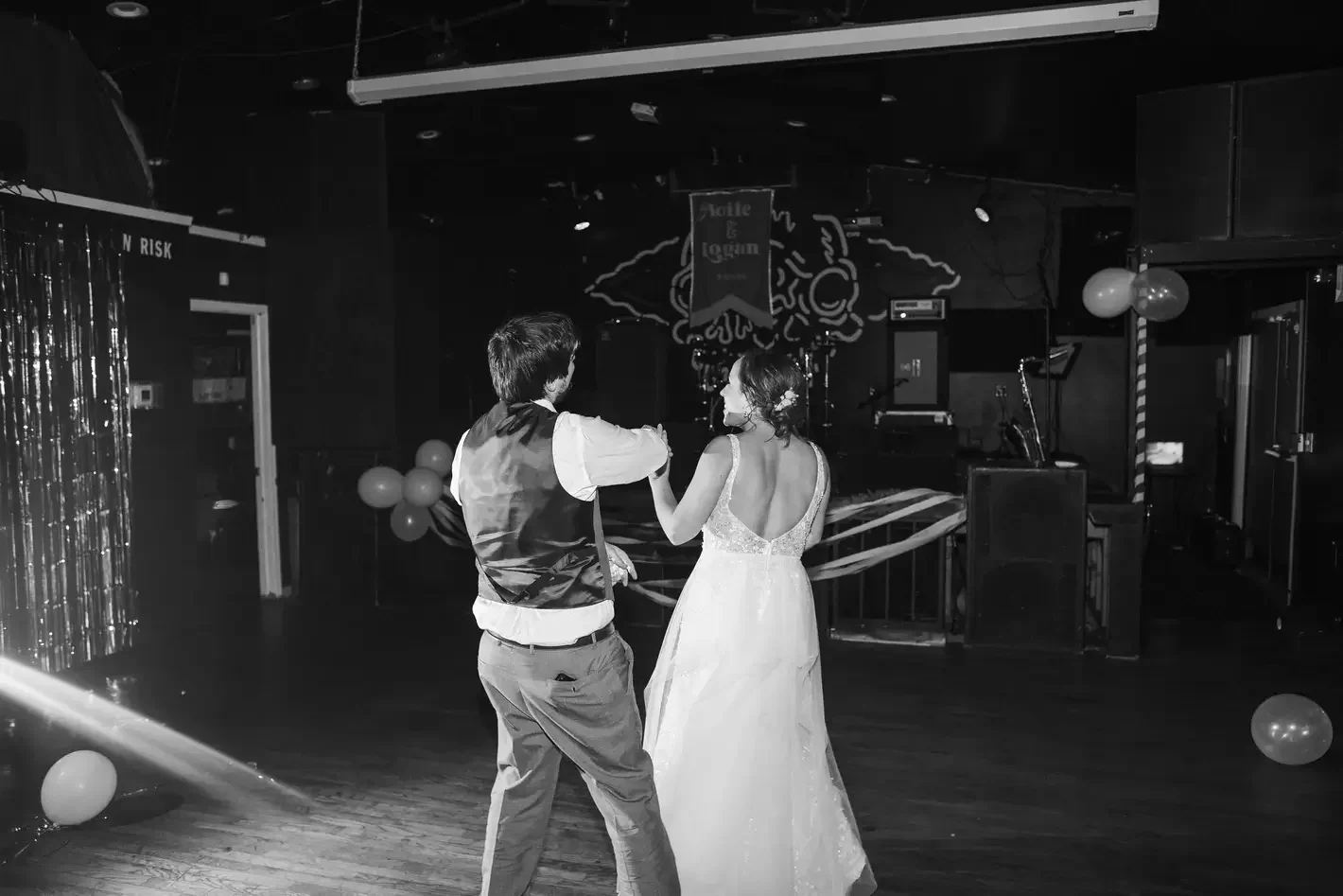 A couple dancing at a wedding reception, with the woman in a white wedding gown and the man in formal attire, in an event space decorated with balloons and balloons, with a stage and musical equipment in the background.