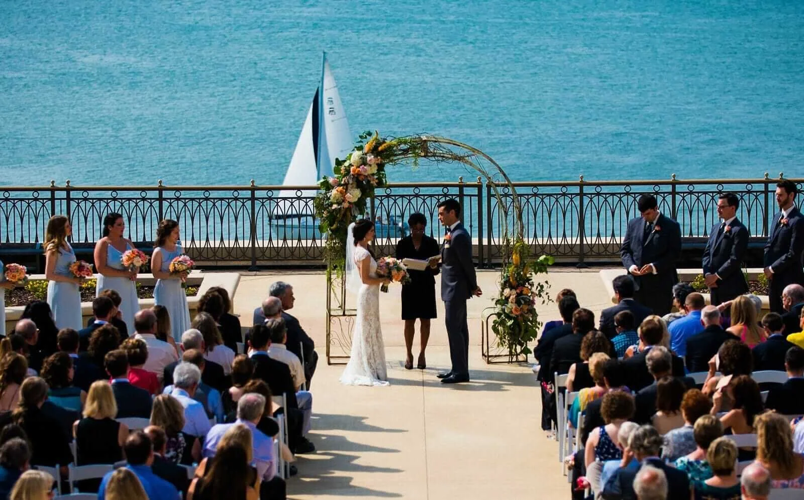 A wedding ceremony taking place outdoors by the water, with the ocean and a sailboat in the background. The bride and groom are standing under a floral arch, exchanging vows, surrounded by bridesmaids and groomsmen. Guests are seated and watching the ceremony.