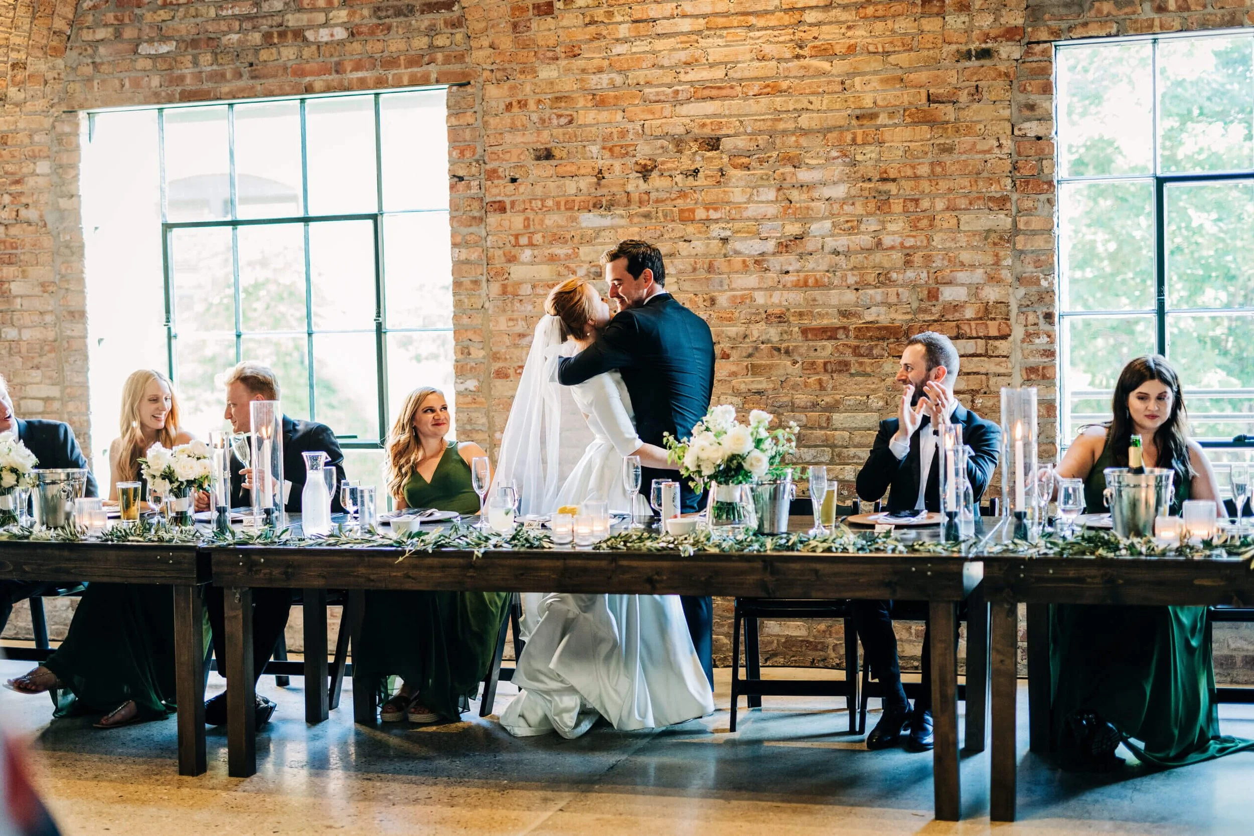 Bride and groom dancing at a wedding reception with guests seated at a table.