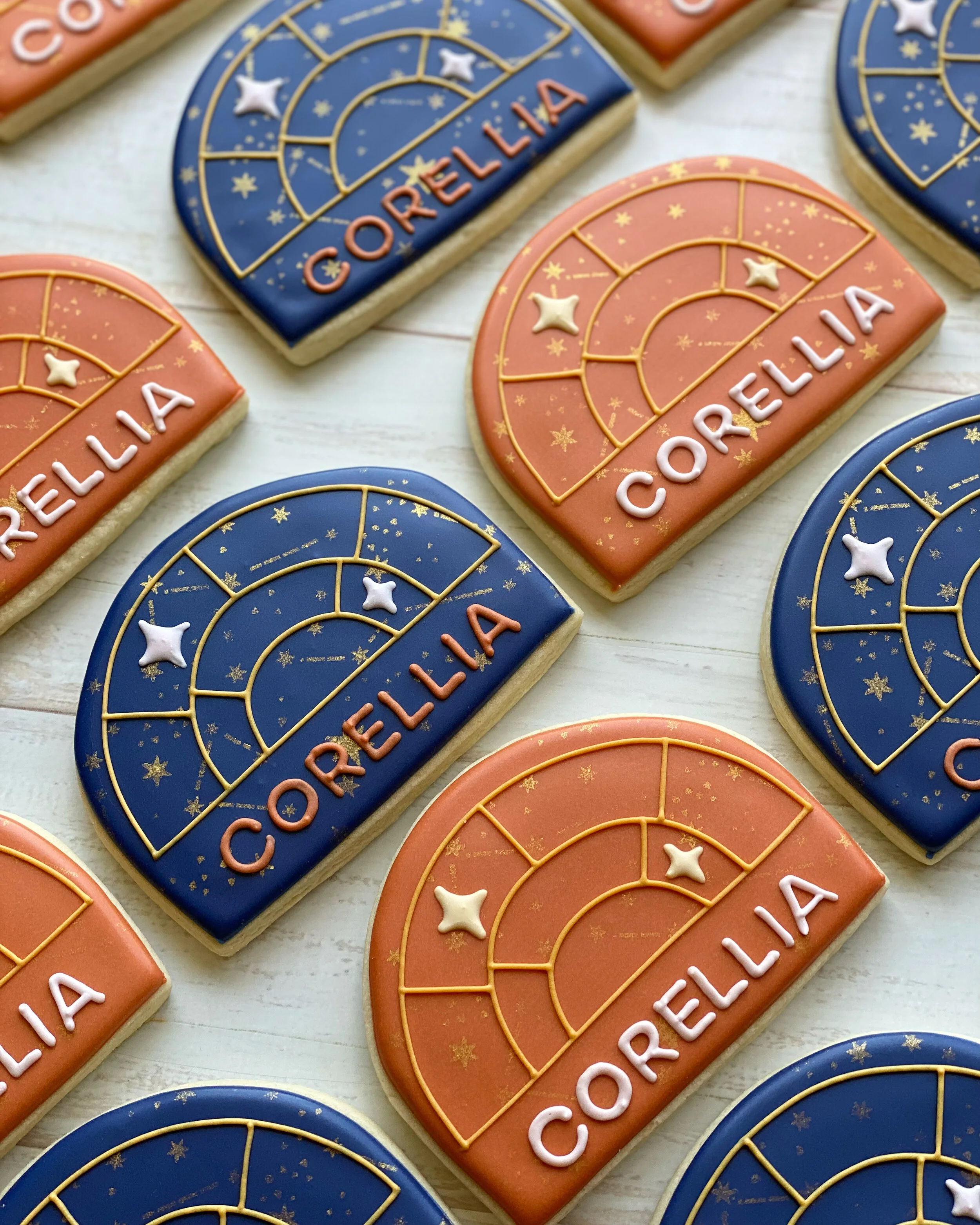 Decorative cookies with a space theme, featuring gold star and planet designs, in blue and peach icing, with the name 'COrELLiA' written in pink and white icing.