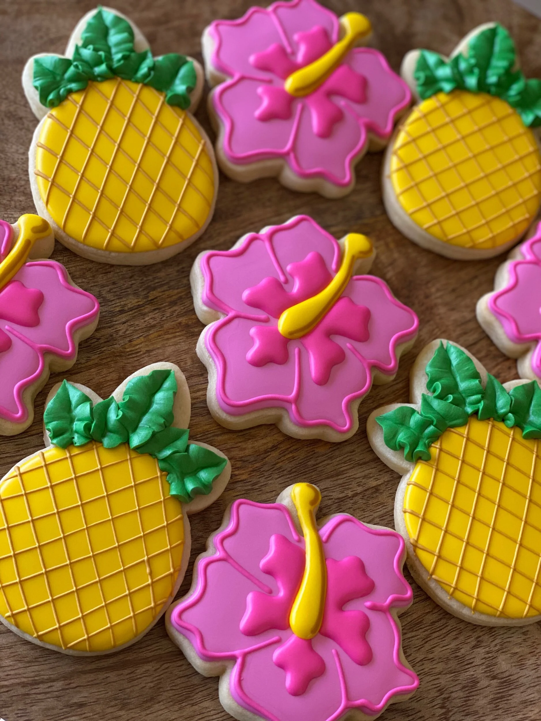 Decorated sugar cookies in tropical and floral shapes, featuring pink, yellow, green, and white icing, arranged on a wooden surface.
