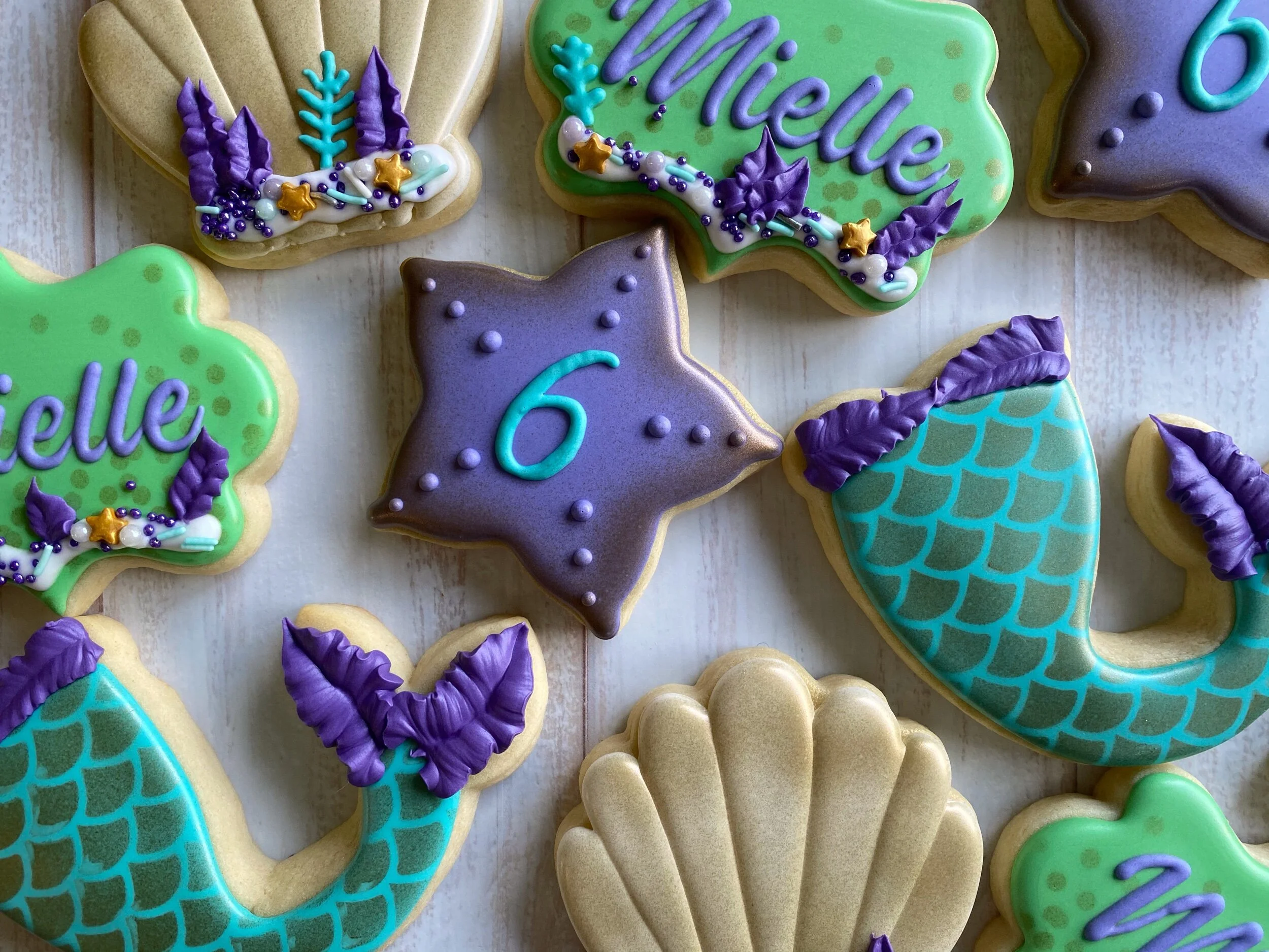 Decorated cookies with various colors and shapes for a birthday celebration, including one shaped like a cat with the number 6, and others with messages and designs in purple, green, blue, and white icing.