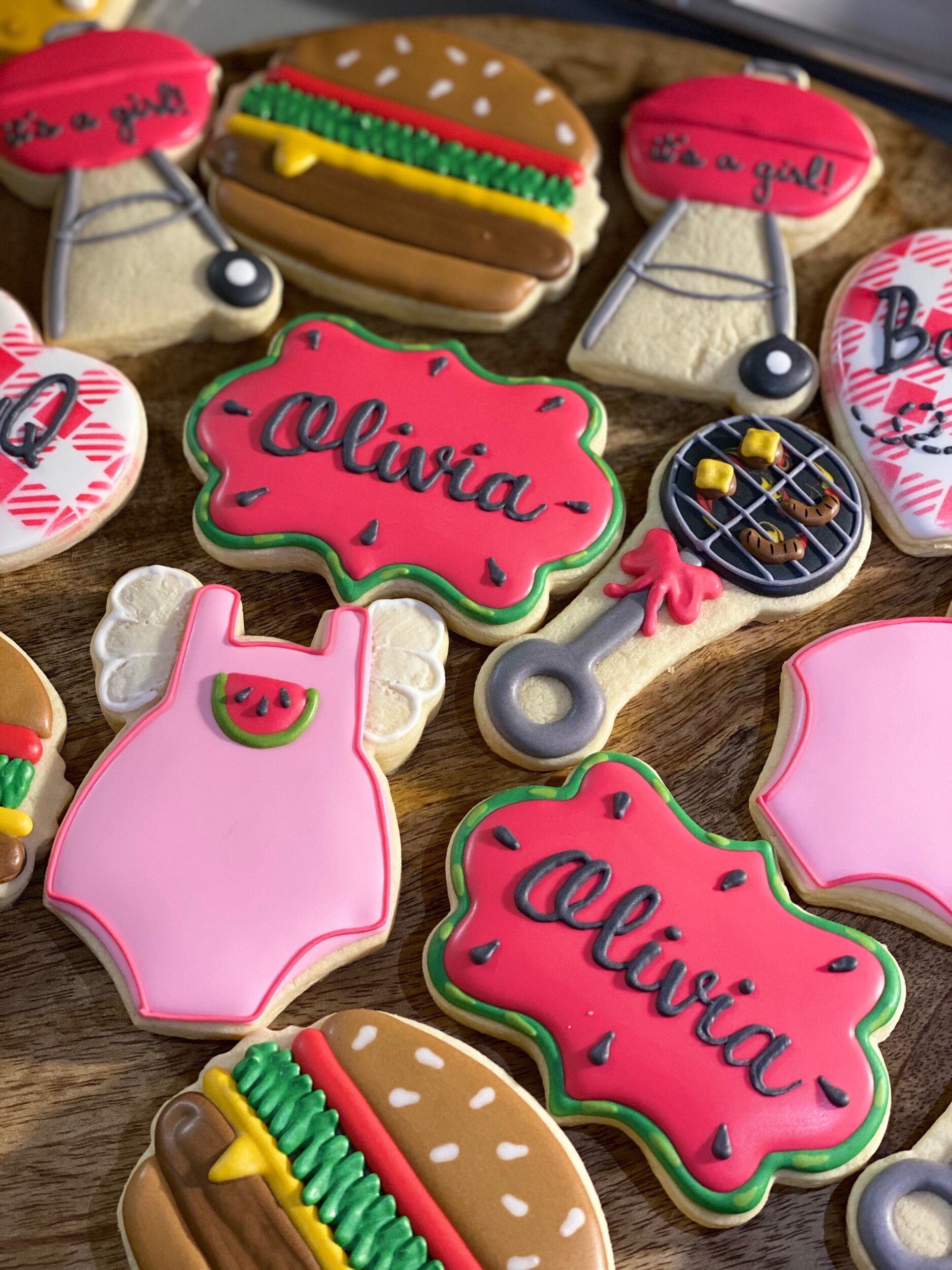 Decorated cookies with phrases like 'it's a girl' and 'Olivia,' shaped and decorated as hot dogs, hamburgers, and other fun shapes, arranged on a wooden surface.