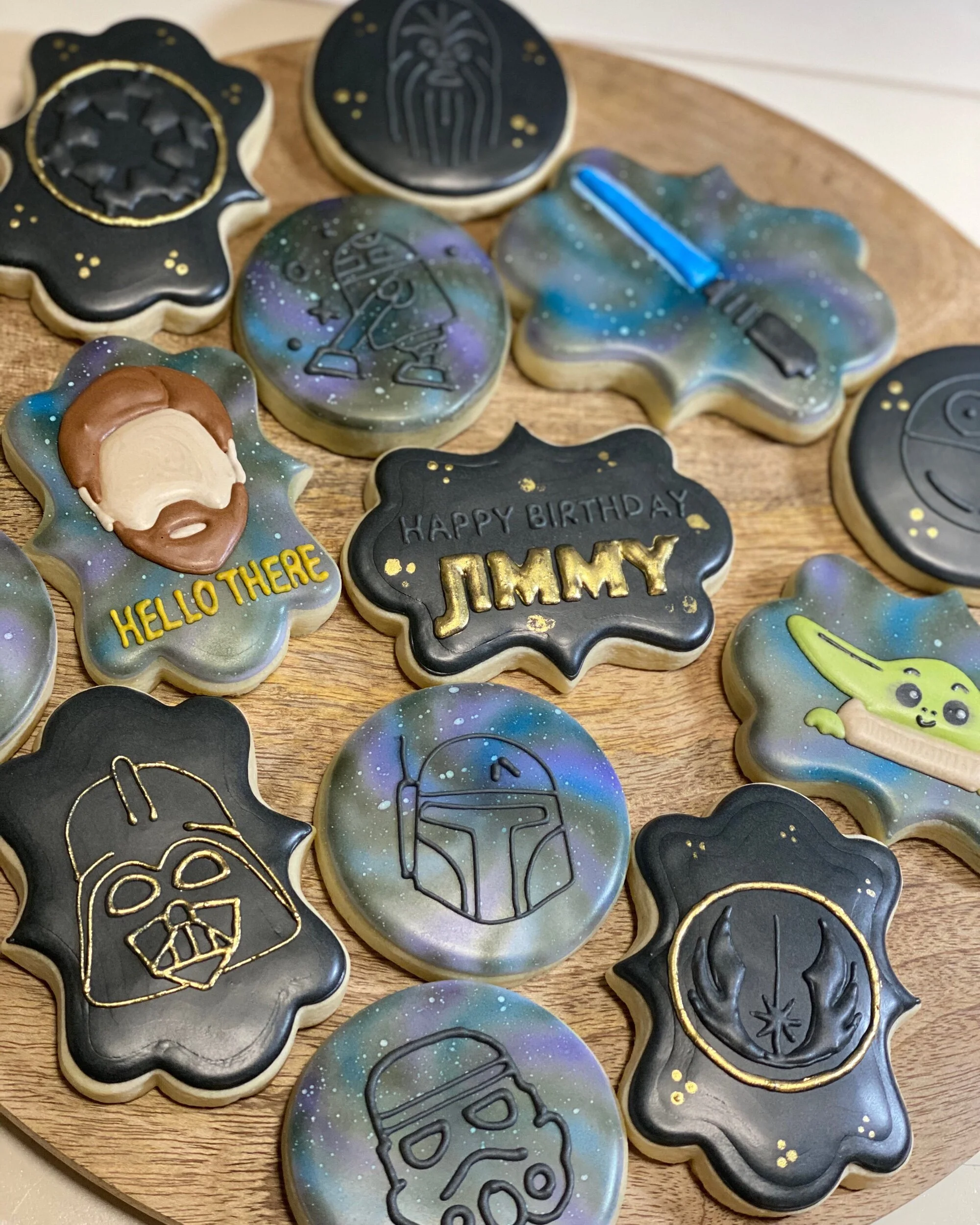 Collection of decorated cookies on a wooden platter featuring space-themed designs and characters from Star Wars, including Yoda, Darth Vader, and R2-D2, with some cookies having black or galaxy-inspired backgrounds and gold accents.