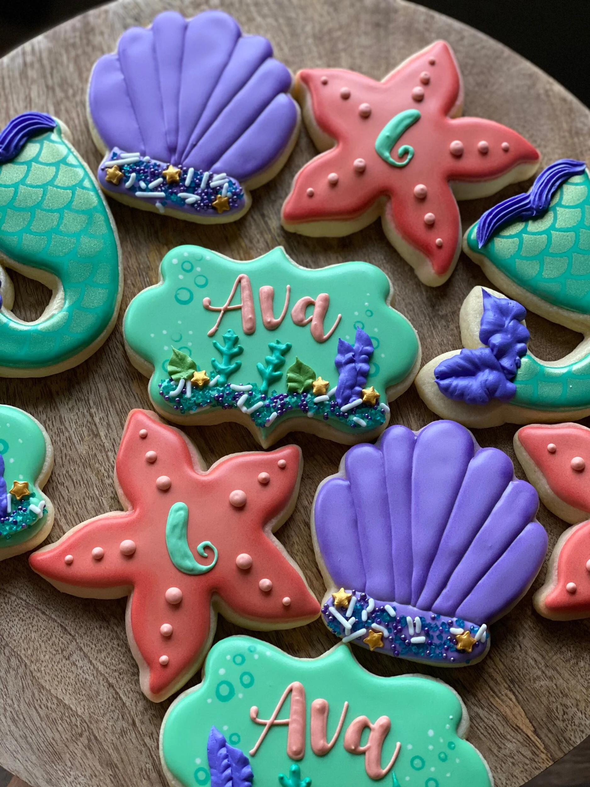 Colorful decorated cookies in sea shell and starfish shapes with personalized messages, arranged on a wooden surface.