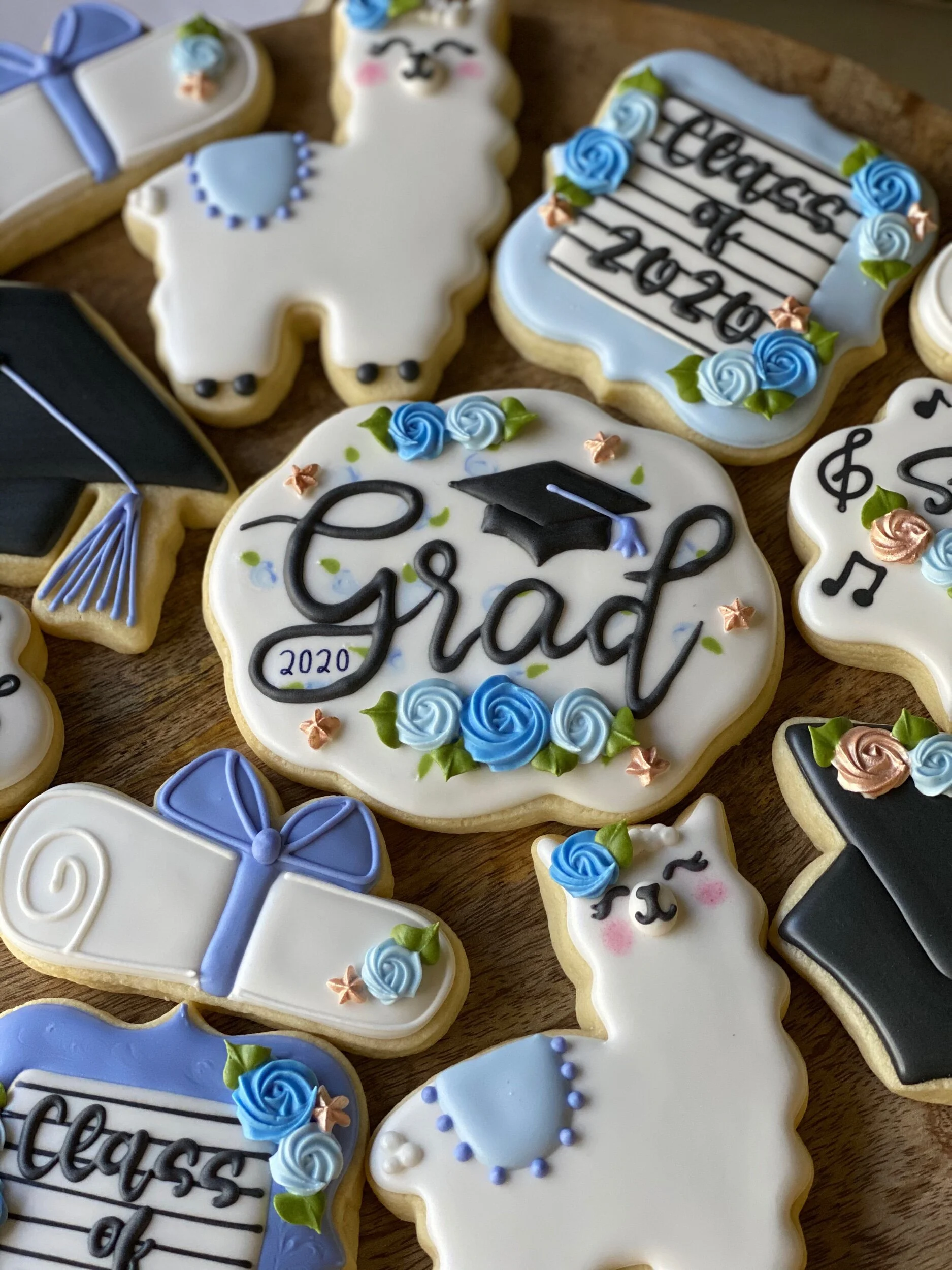 Decorative cookies shaped like graduation caps, diplomas, and graduate faces, decorated with blue, white, black, and pink icing, arranged on a wooden surface.