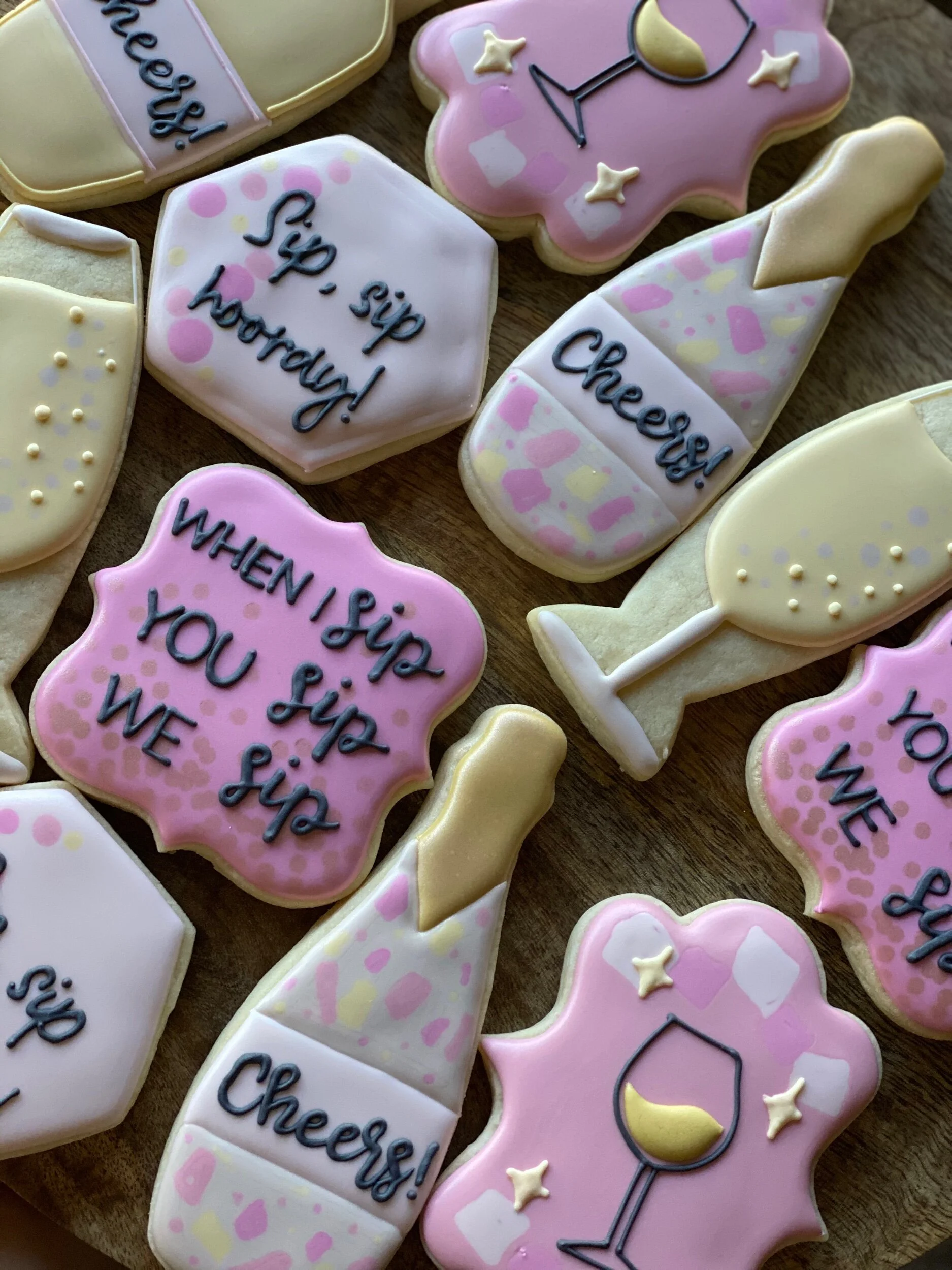 Sugar cookies decorated with pink, white, and yellow icing. Some cookies have messages like "sip sip hooray!" and "when sip you sip." Others are decorated with wine glasses, champagne bottles, and star accents.