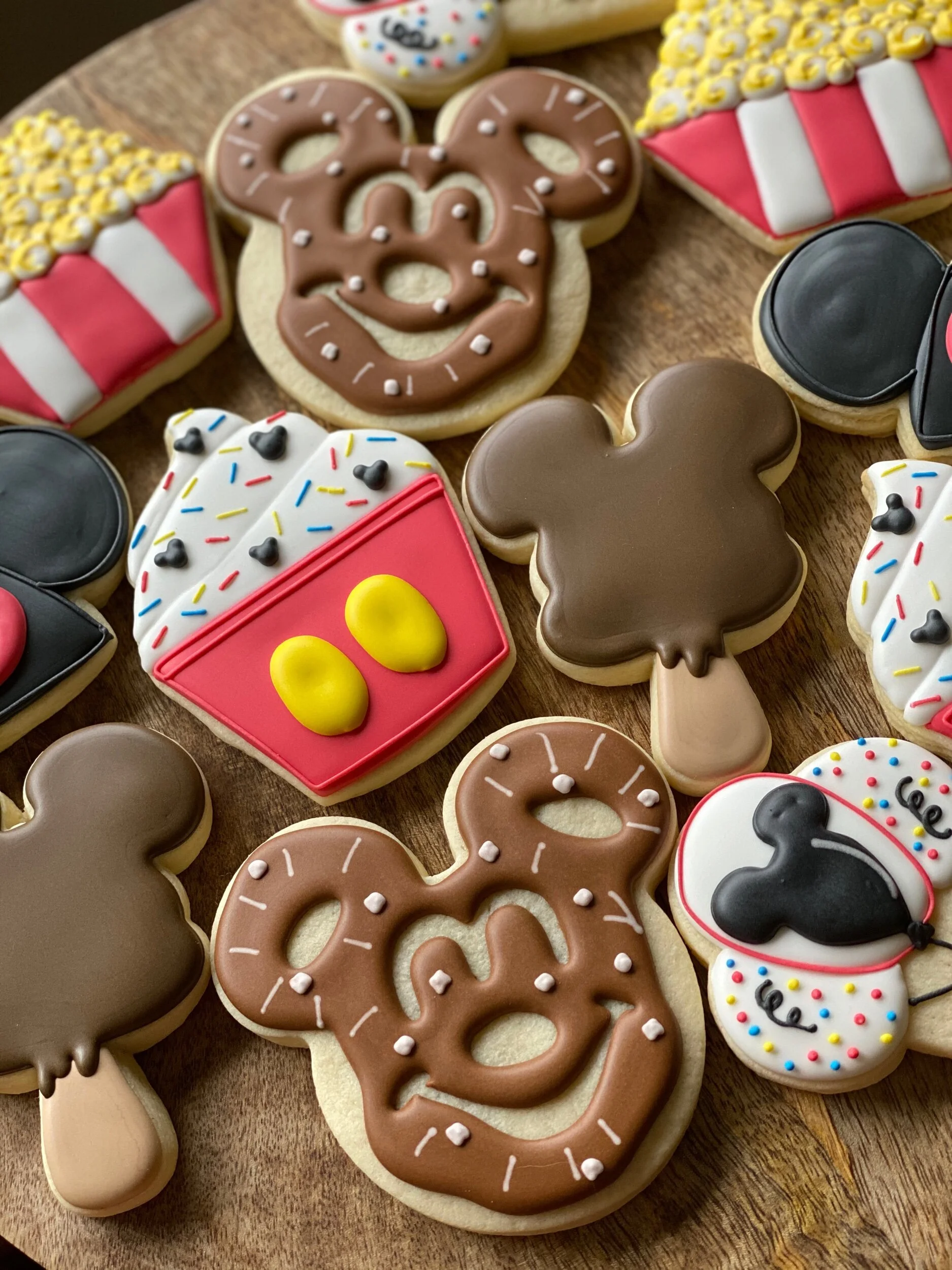 Assorted decorated cookies themed for a celebration, including Mickey Mouse, pretzels with brown and white icing, popcorn buckets with yellow and blue sprinkles, and other colorful designs, all on a wooden surface.