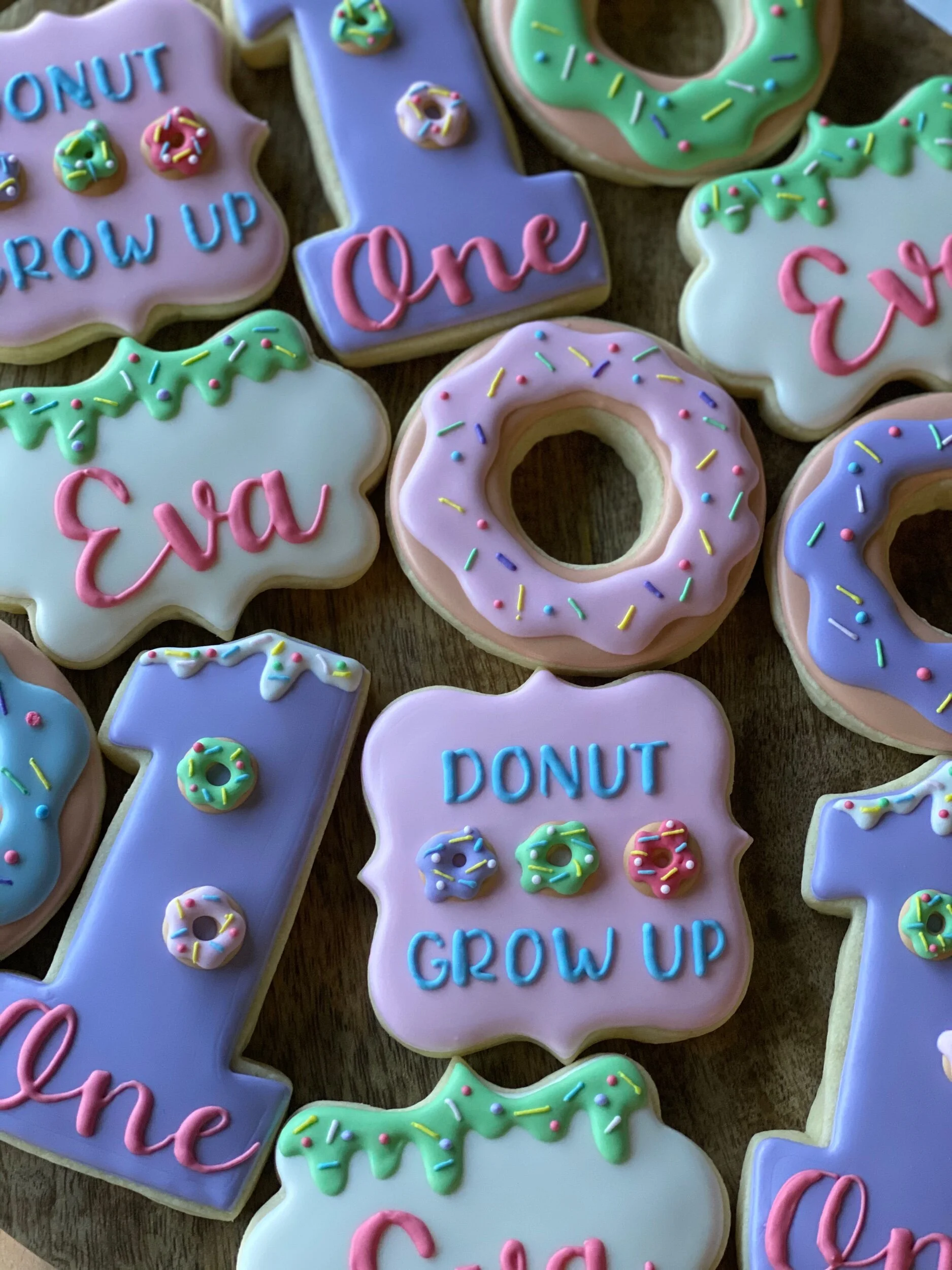Colorfully decorated cookies with messages like 'Grow Up,' 'One,' and 'Donut Grow Up' in pastel icing, sprinkles, and small donut-shaped decorations on a wooden surface.