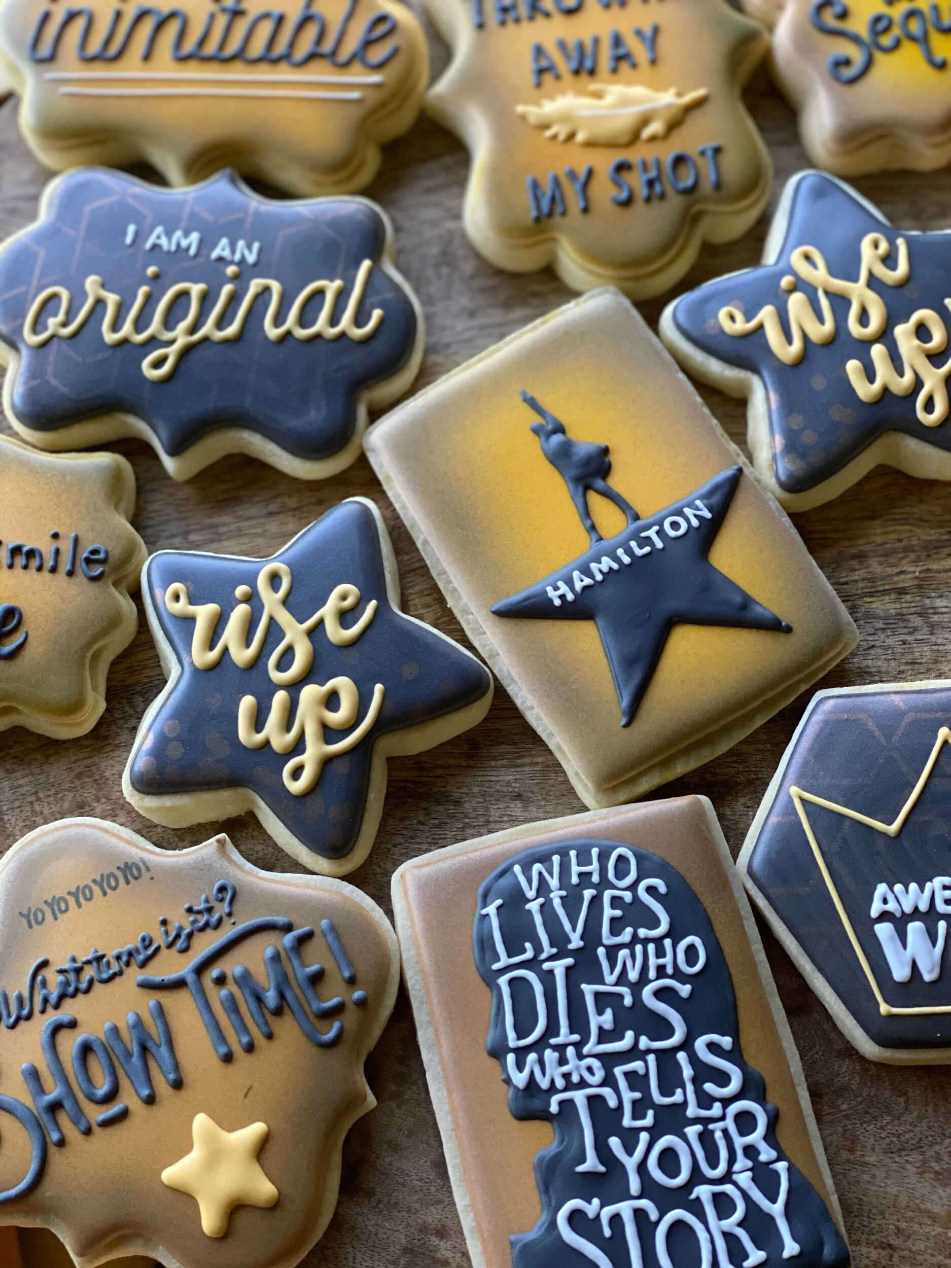 Assorted decorated cookies with motivational and humorous phrases such as 'I am an original,' 'talks away my shot,' and 'who tells your story,' featuring stars, a person doing yoga, and abstract designs.