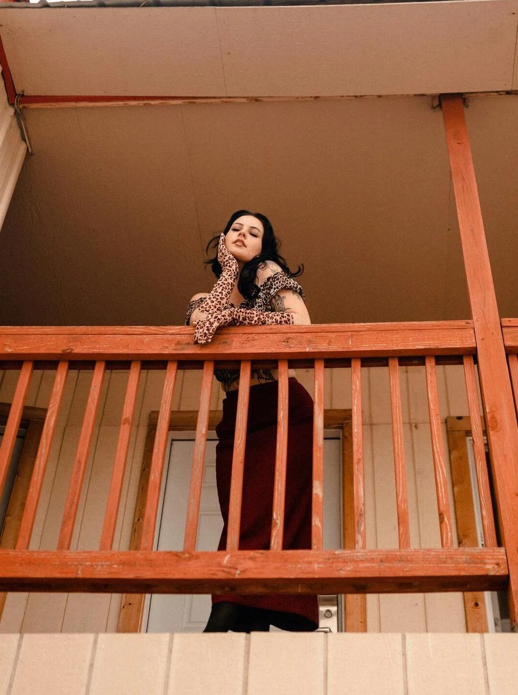 A woman with dark hair and tattoos posing on a wooden balcony, wearing a leopard print long gloves and top, with dark red pants, striking a confident pose.
