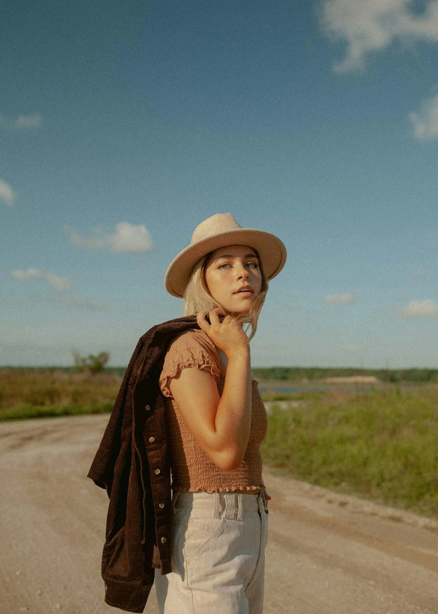 A young woman with blonde hair wearing a tan hat, brown t-shirt, and light blue jeans, standing outdoors on a dirt road with a grassy field and blue sky with a few clouds in the background.