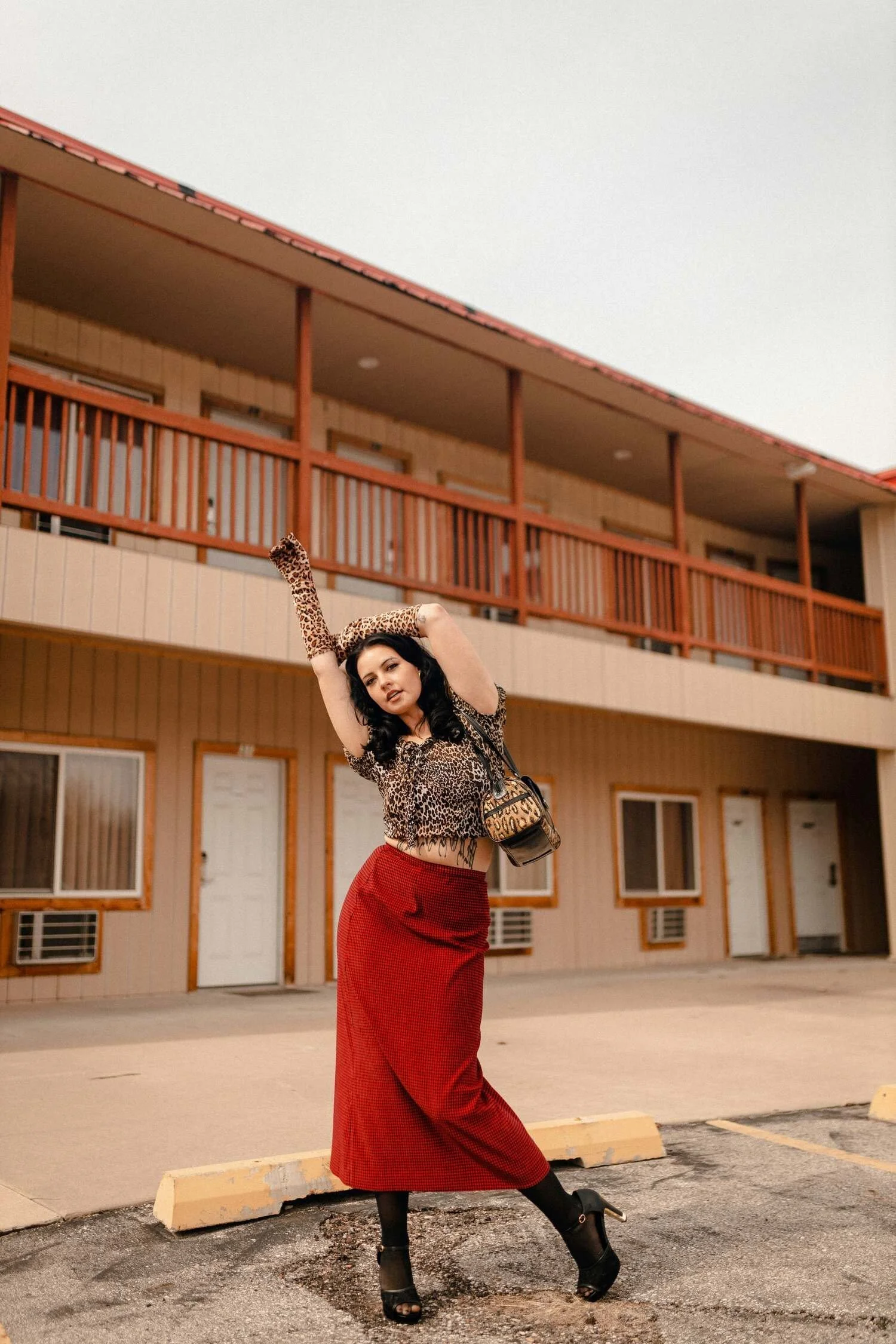 A woman with dark, curly hair and wearing leopard print gloves, a leopard print top, and a long red skirt, posing with one arm raised and looking at the camera. She is standing in an empty parking lot in front of a two-story motel with a balcony and wooden railing.