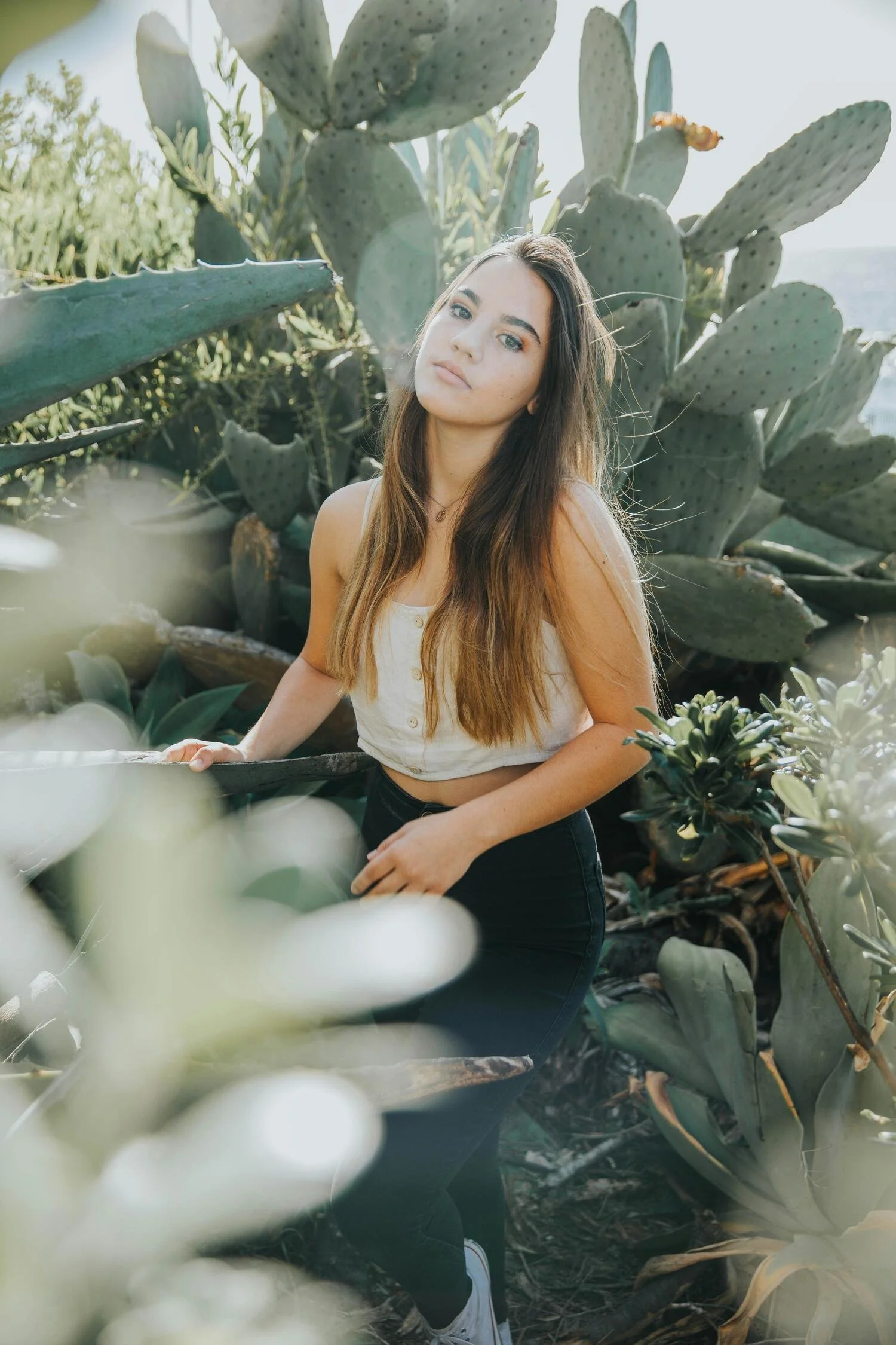 Young woman with long brown hair and light skin standing among large green cactus plants outdoors.