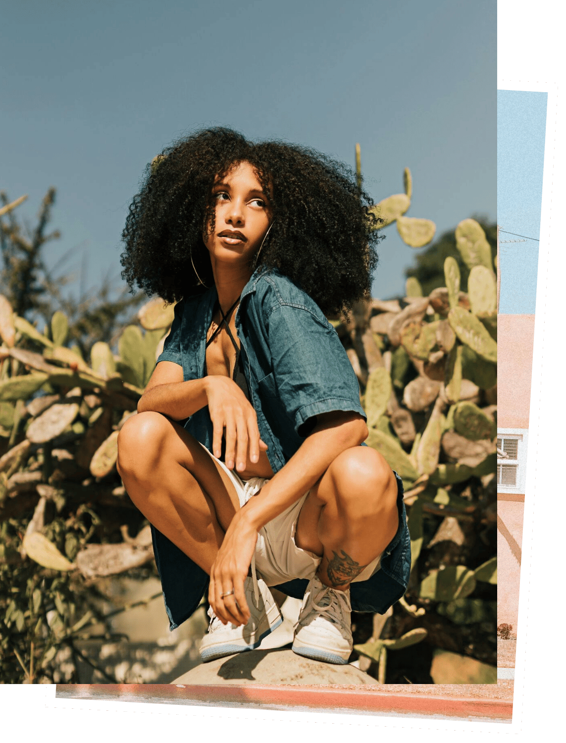 A young woman with curly hair squatting outdoors among cactus plants, wearing a denim shirt, beige shorts, and white sneakers, with a serious expression.
