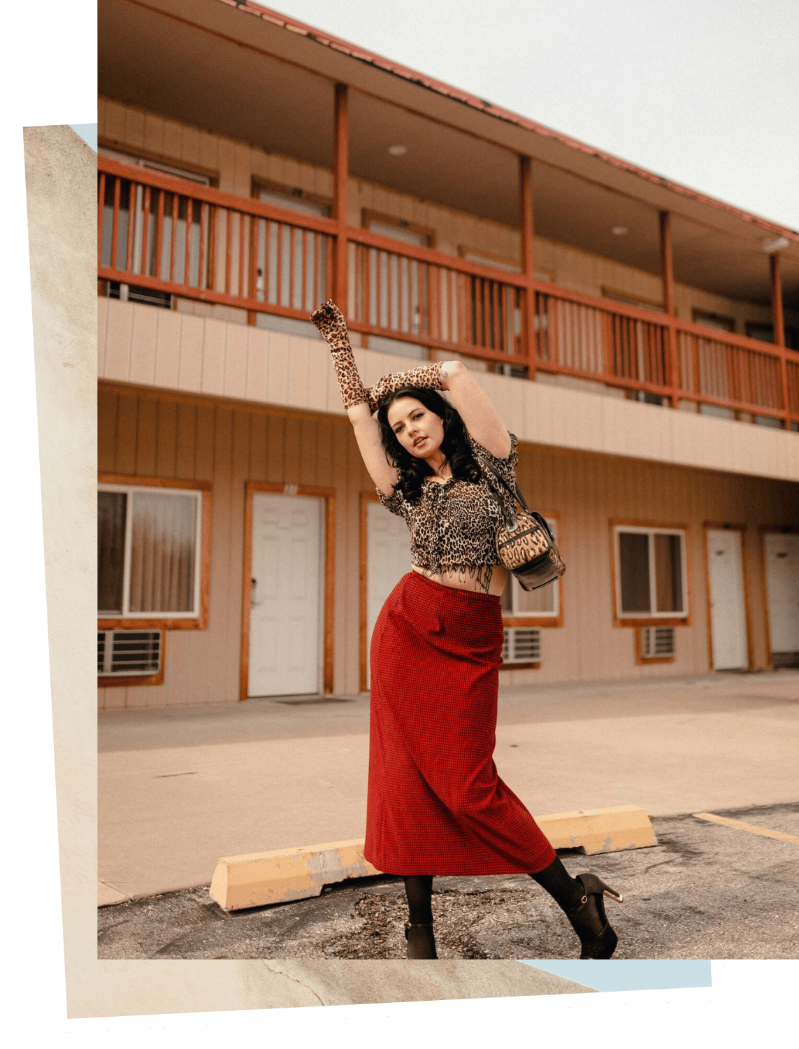 A woman wearing a leopard print top, red skirt, and leopard gloves posing with her arms raised in front of a two-story motel with wooden siding and a balcony.