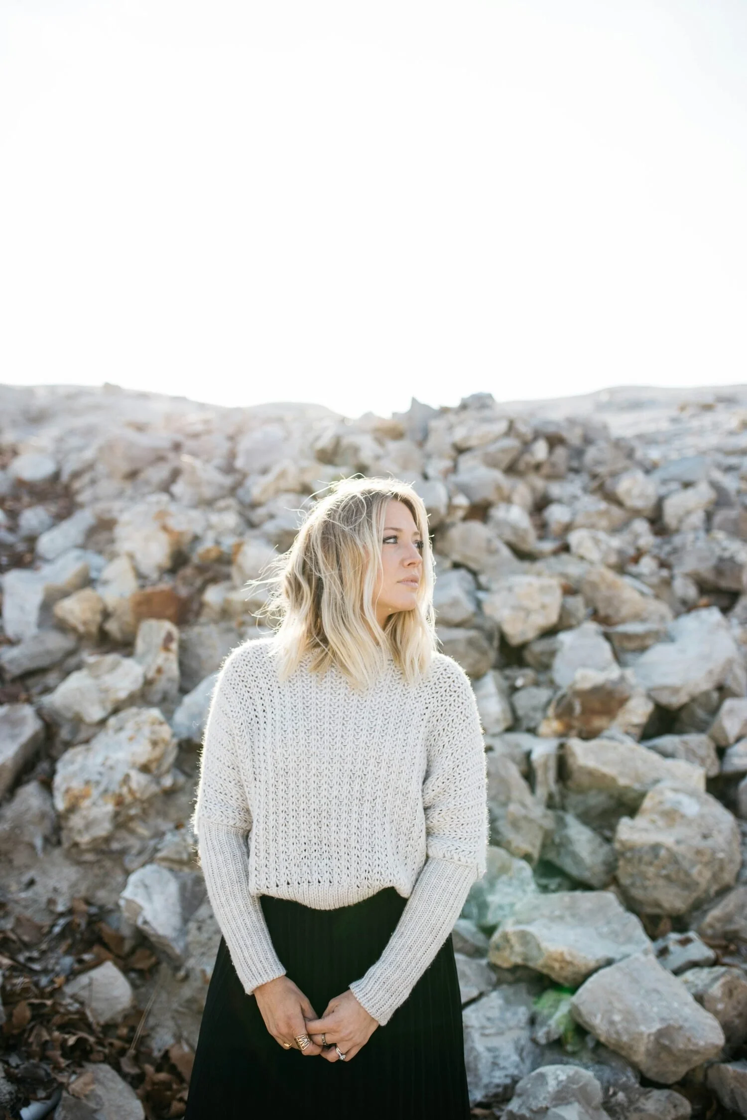 A woman with blonde hair wearing a cream sweater and black skirt stands in front of a rocky terrain with rocks and stones in the background. The sky appears bright and clear.
