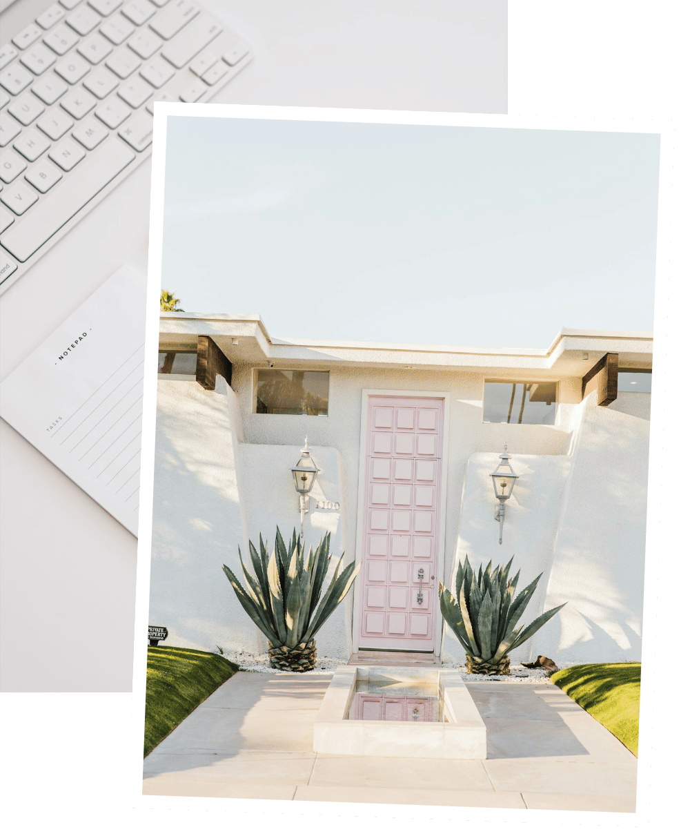Front entrance of a modern white house with a pink door, two large agave plants, and decorative outdoor lamps. Outside the house, there is a built-in rectangular water feature and a well-manicured lawn.