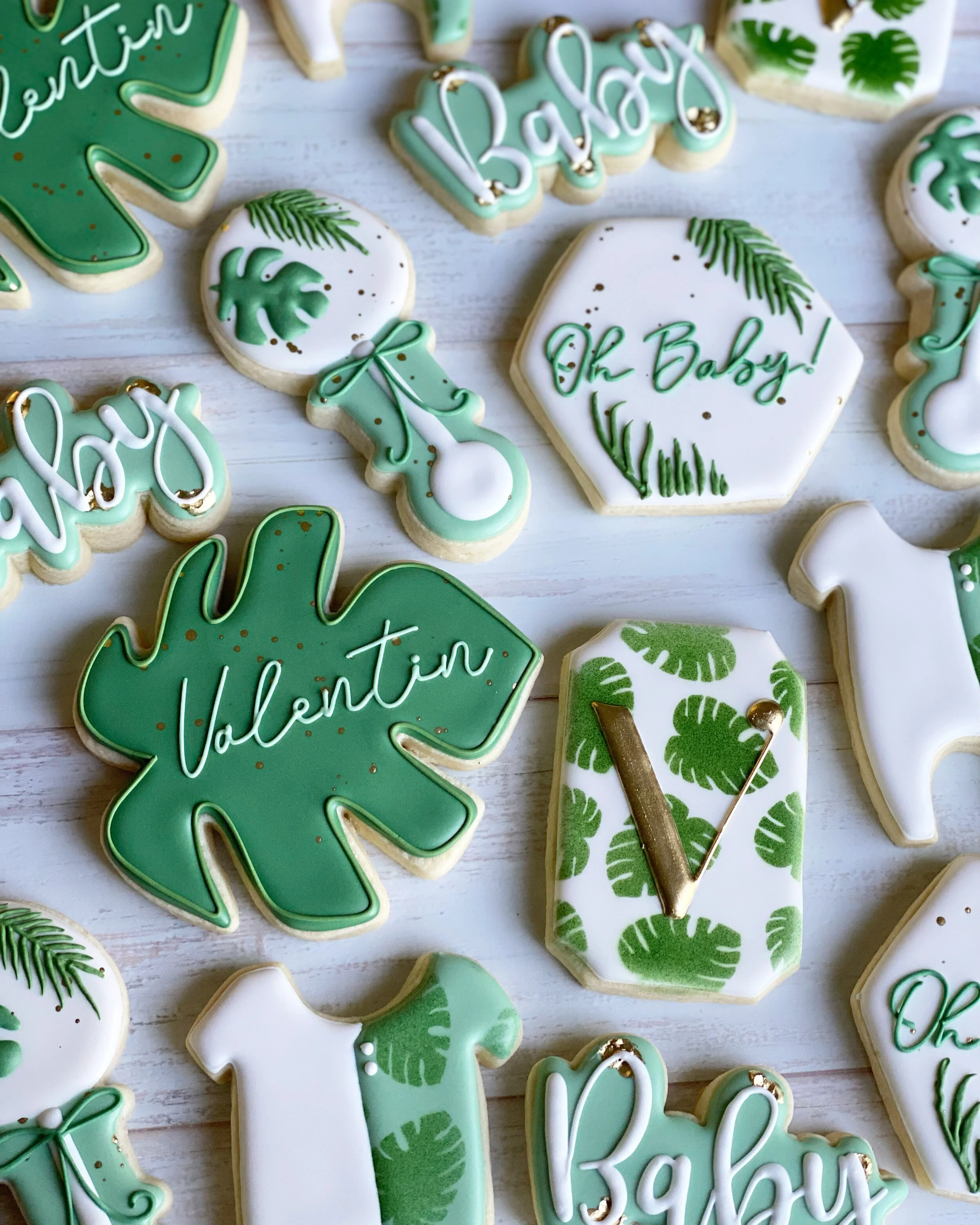 Assorted decorated cookies with a green, white, and gold color theme for a baby shower. The cookies feature designs such as the words 'Baby' and 'Valentin,' a baby onesie, a pacifier, and tropical leaves.