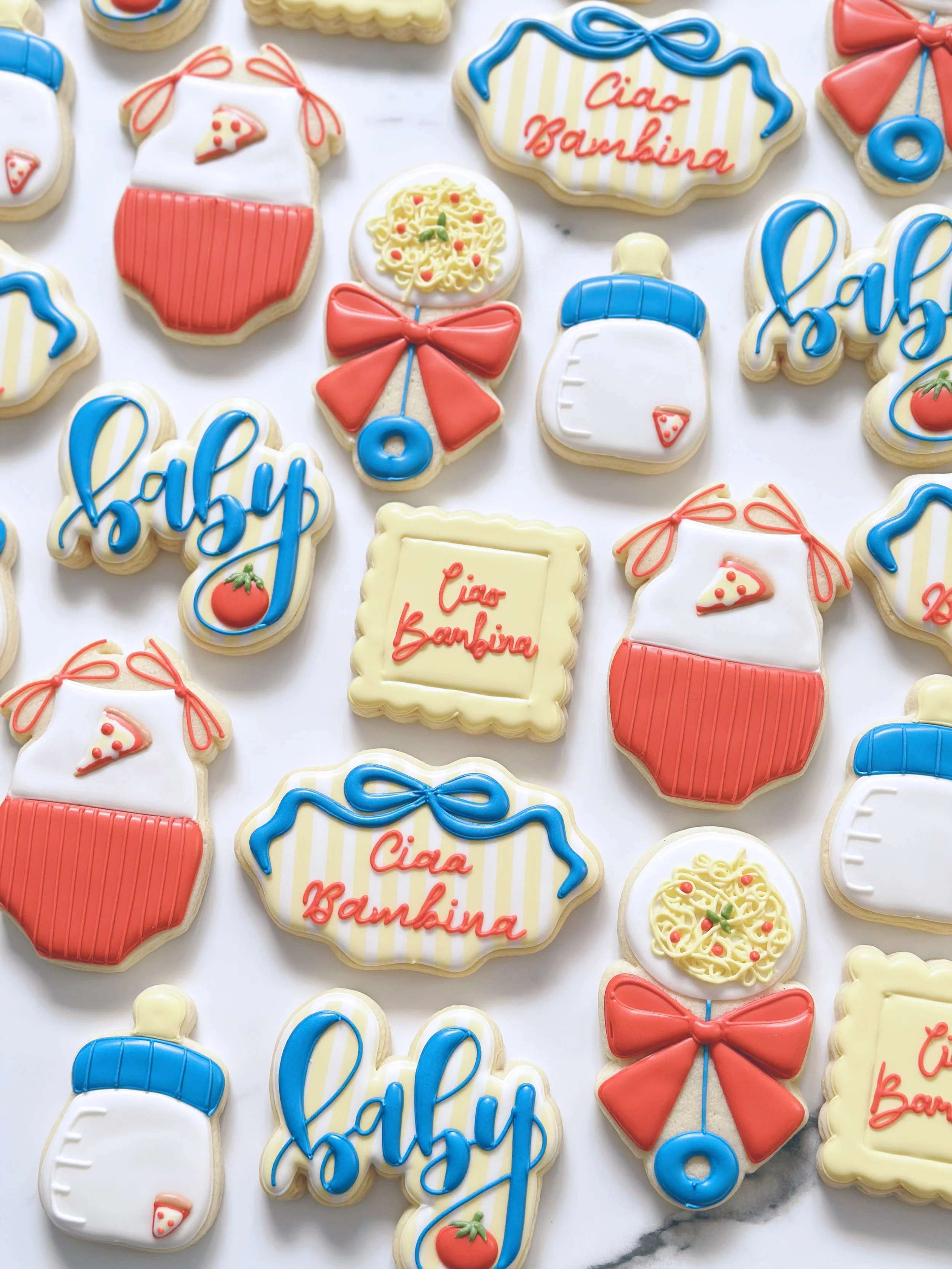 Colorful cookies decorated with pasta, pizza, tomato, and text messages like 'Ciao Bambina' and 'Baby' in blue and red icing, arranged on a white marble surface.