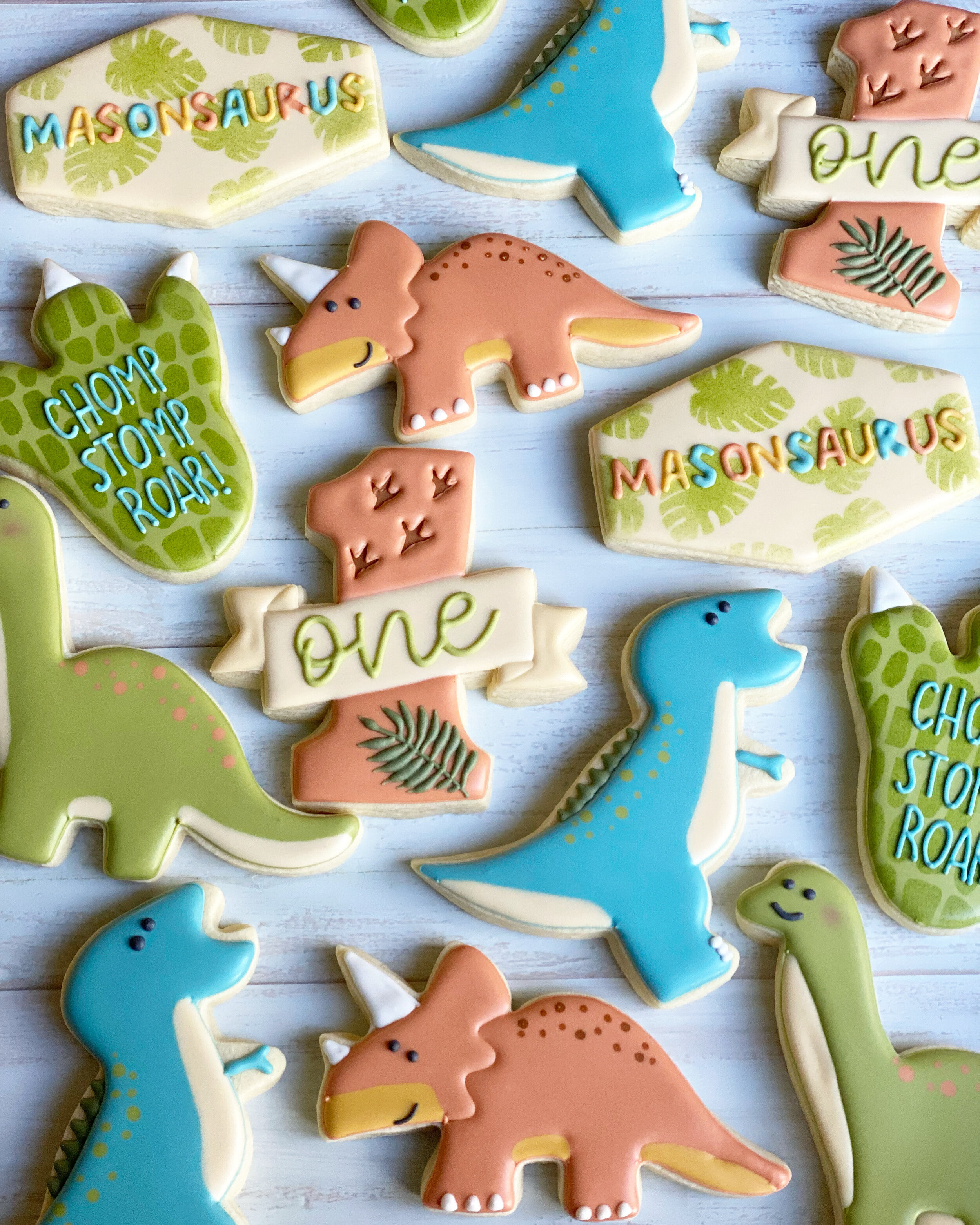A collection of dinosaur-shaped cookies decorated with colorful icing including designs with the words "MASONSAURUS", "CHOMP CHOMP ROAR!", and "one" on some of them. The cookies are arranged on a light wooden surface.