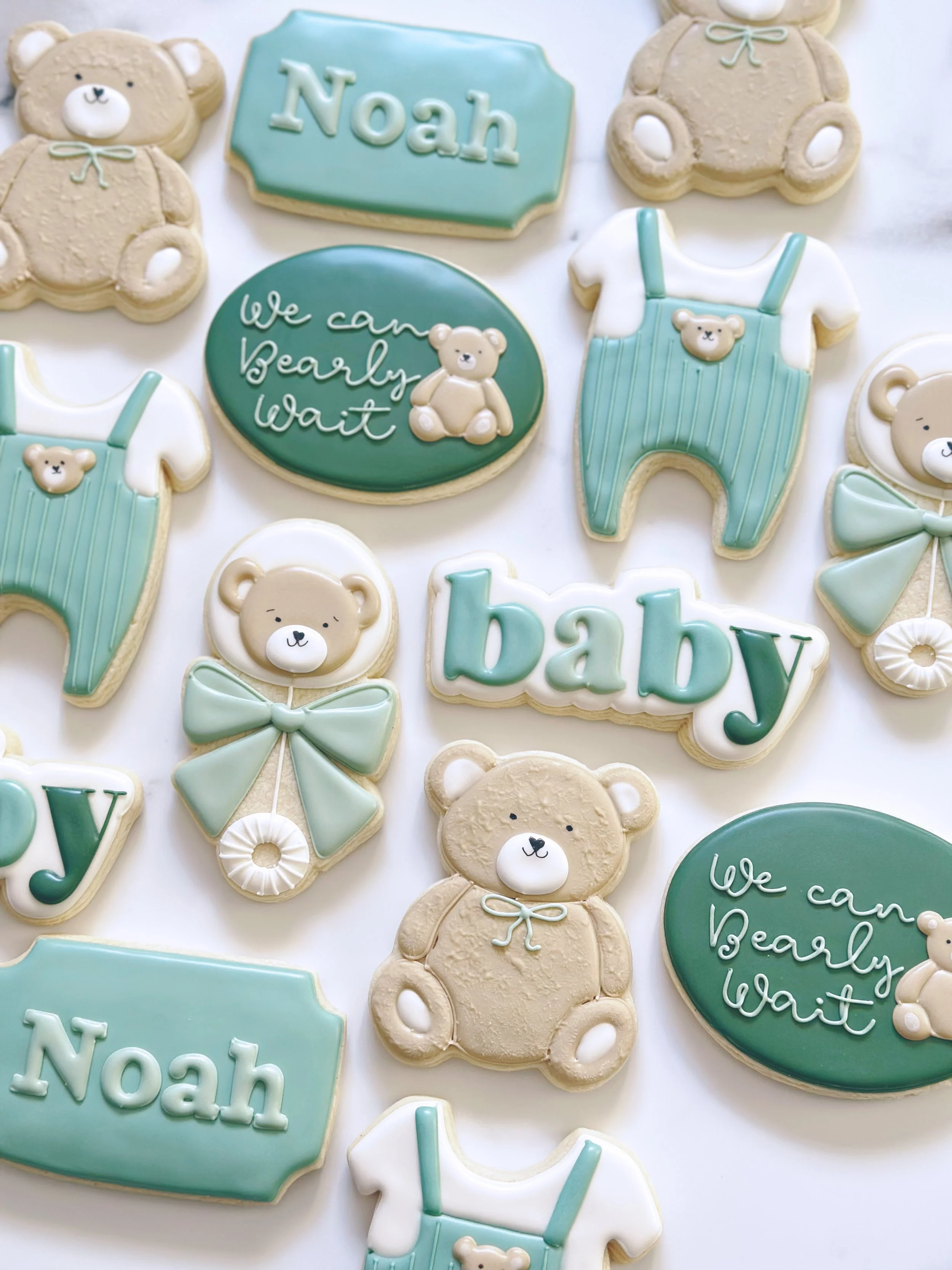 Baby shower cookies decorated with teddy bear designs, baby clothing, and messages like "Noah" and "We can bearly wait" in pastel blue, white, and cream colors.