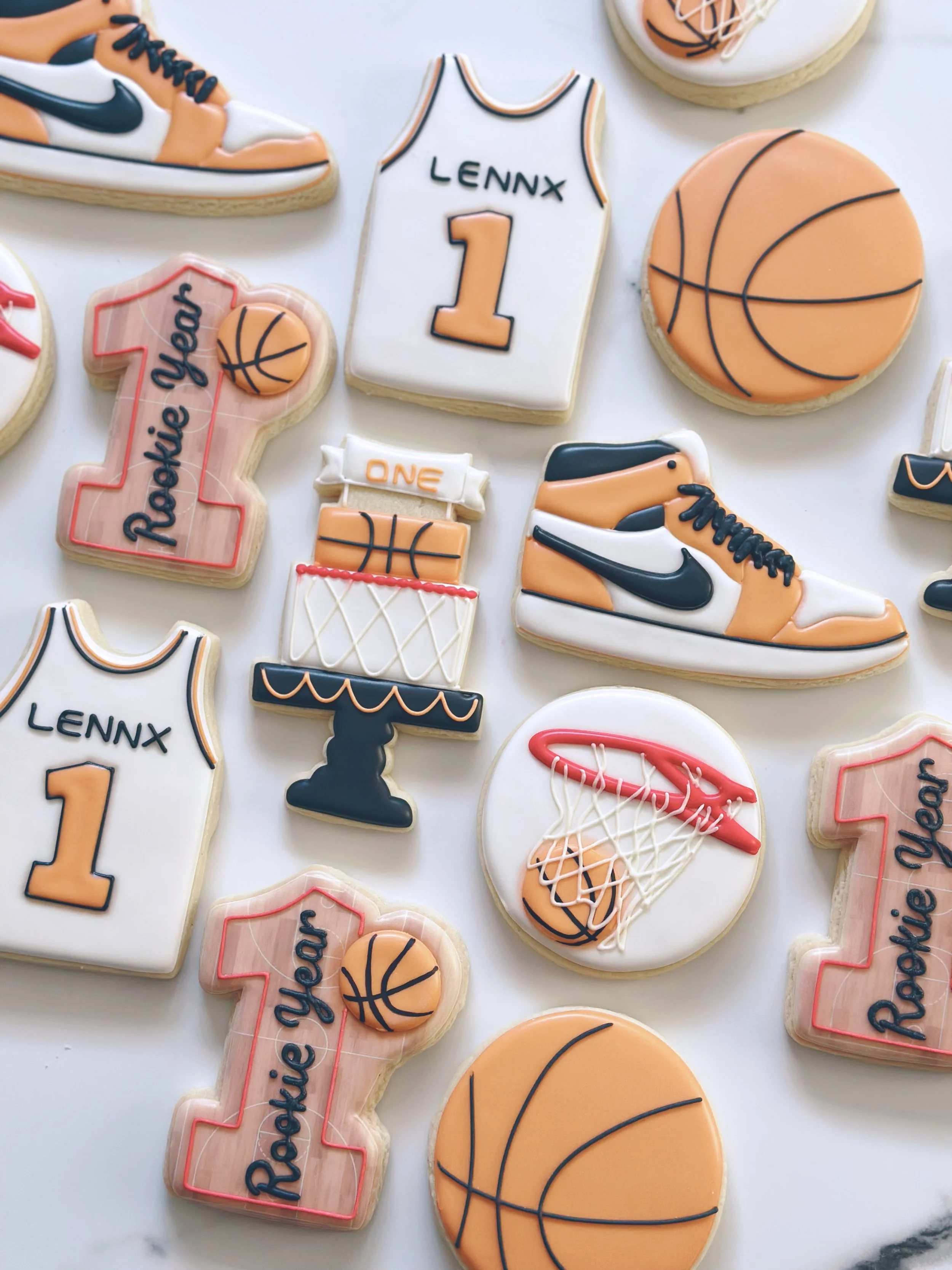 Decorative cookies with basketball and sport-themed designs, including a jersey with the name 'Lennx' and the number '1', a basketball, a basketball hoop, and text 'Rosie you' on a pink and white cookie.