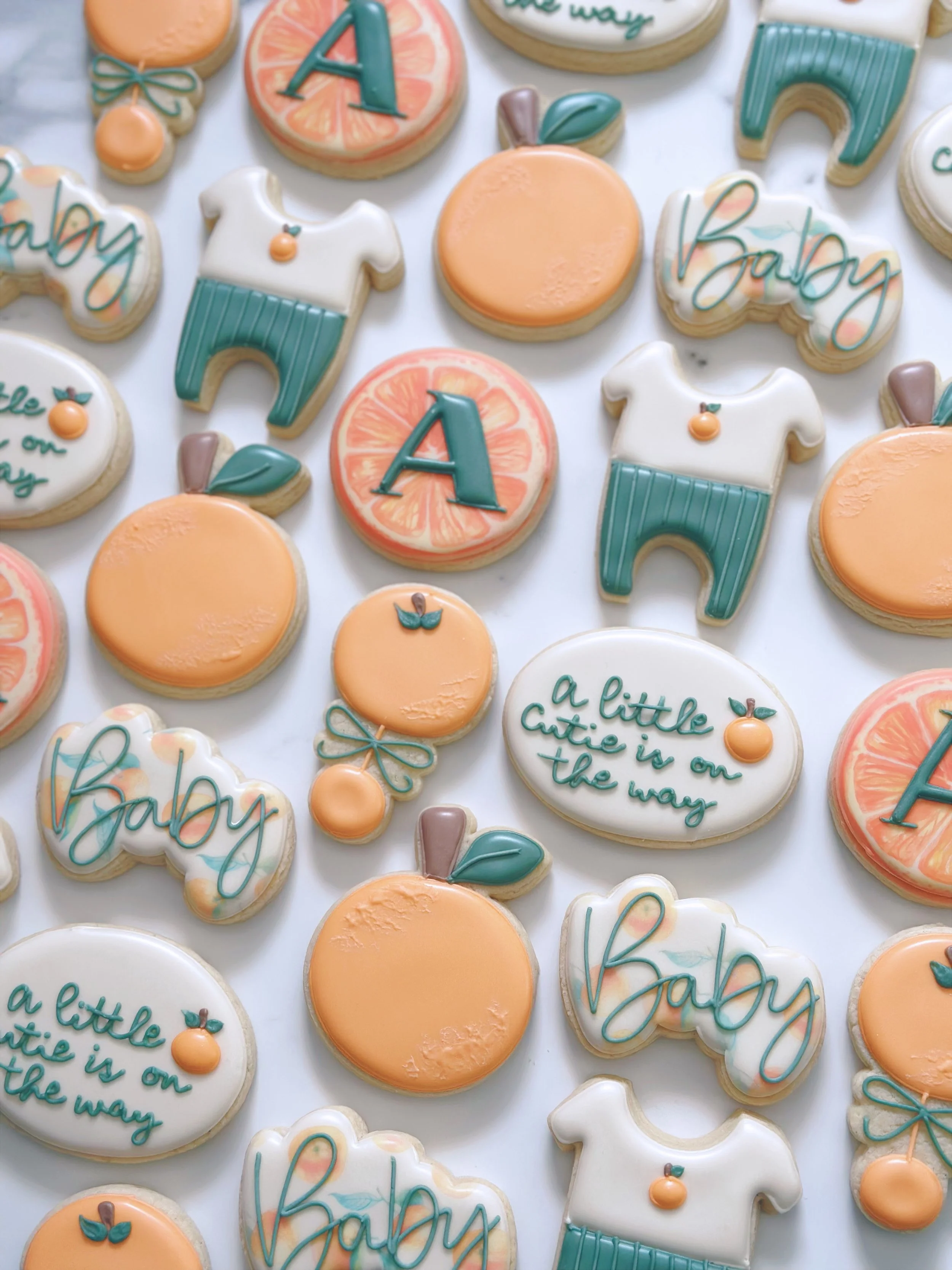 Assorted decorated cookies with baby-themed designs including the word "baby," oranges, and baby clothes, in pastel colors of peach, white, and teal.