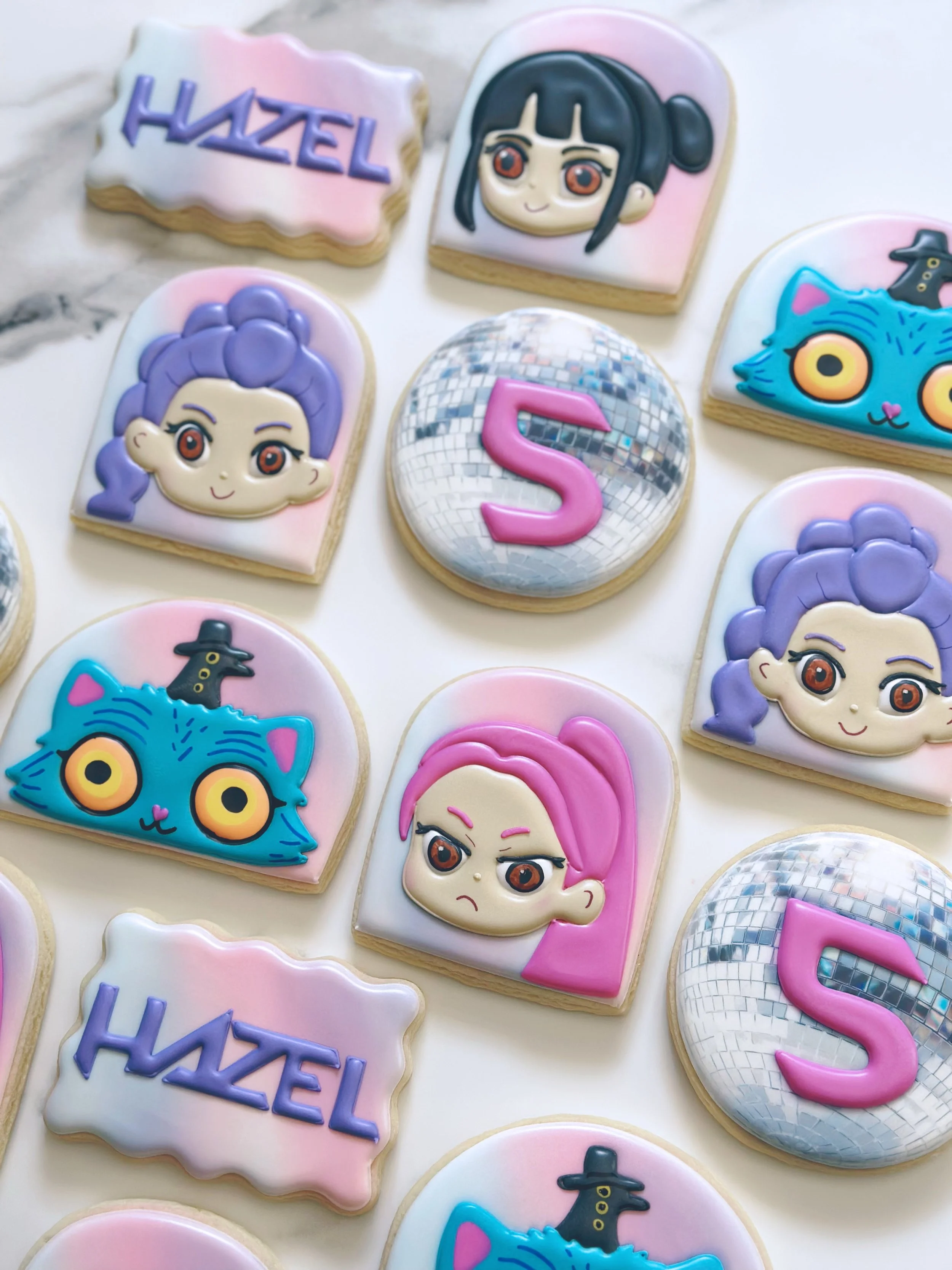 Decorated sugar cookies with cartoon characters, a disco ball, a pink number 5, and the word 'Hazel' in purple letters.