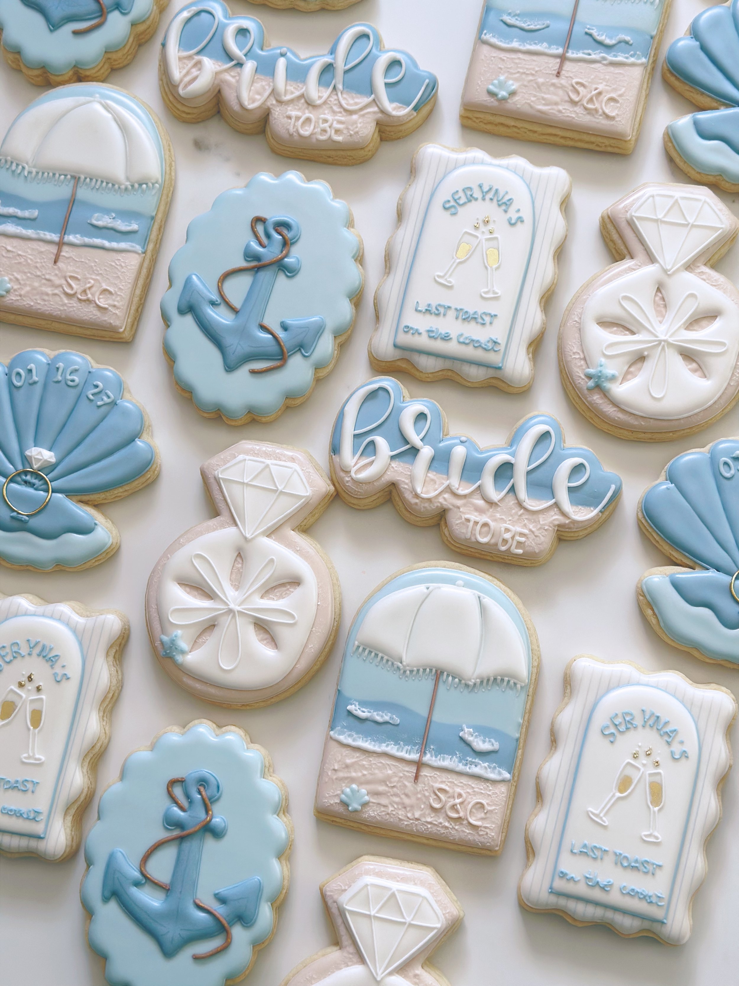 Assorted beach-themed decorated cookies with pastel blue, white, and pink icing, featuring designs such as umbrellas, anchors, diamonds, clouds, and messages like "Bride To Be" and "Serena's Last Toast on the Coast".
