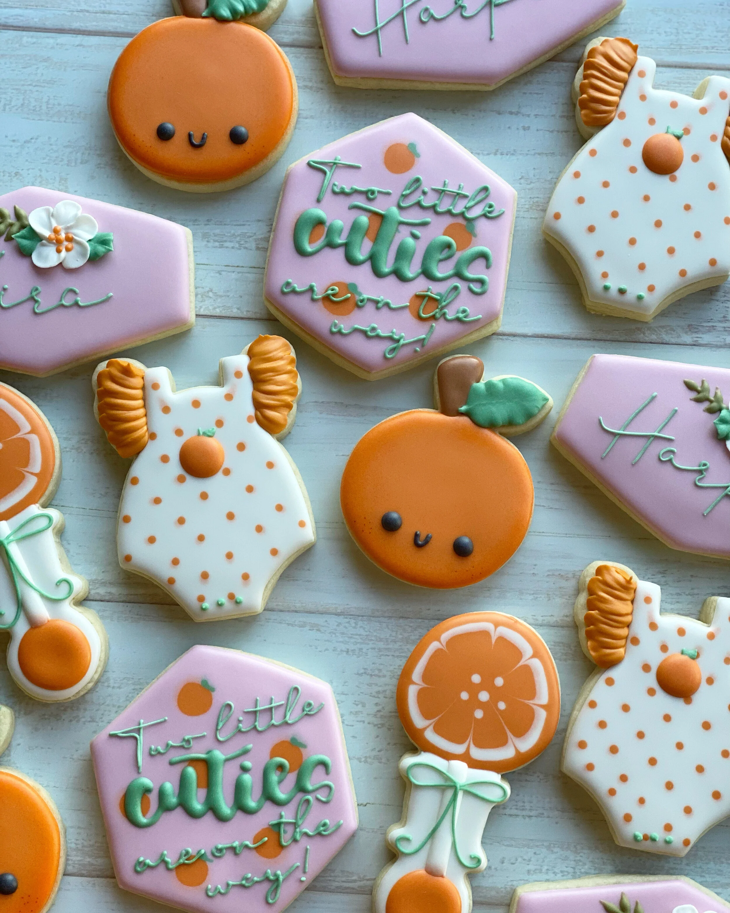 Decorated sugar cookies with orange, pink, white, and green icing. The cookies feature flowers, oranges, and the phrase "Two little cuties over the way!" in colorful icing. Some cookies are shaped like oranges and a girl wearing a dress with pigtails
