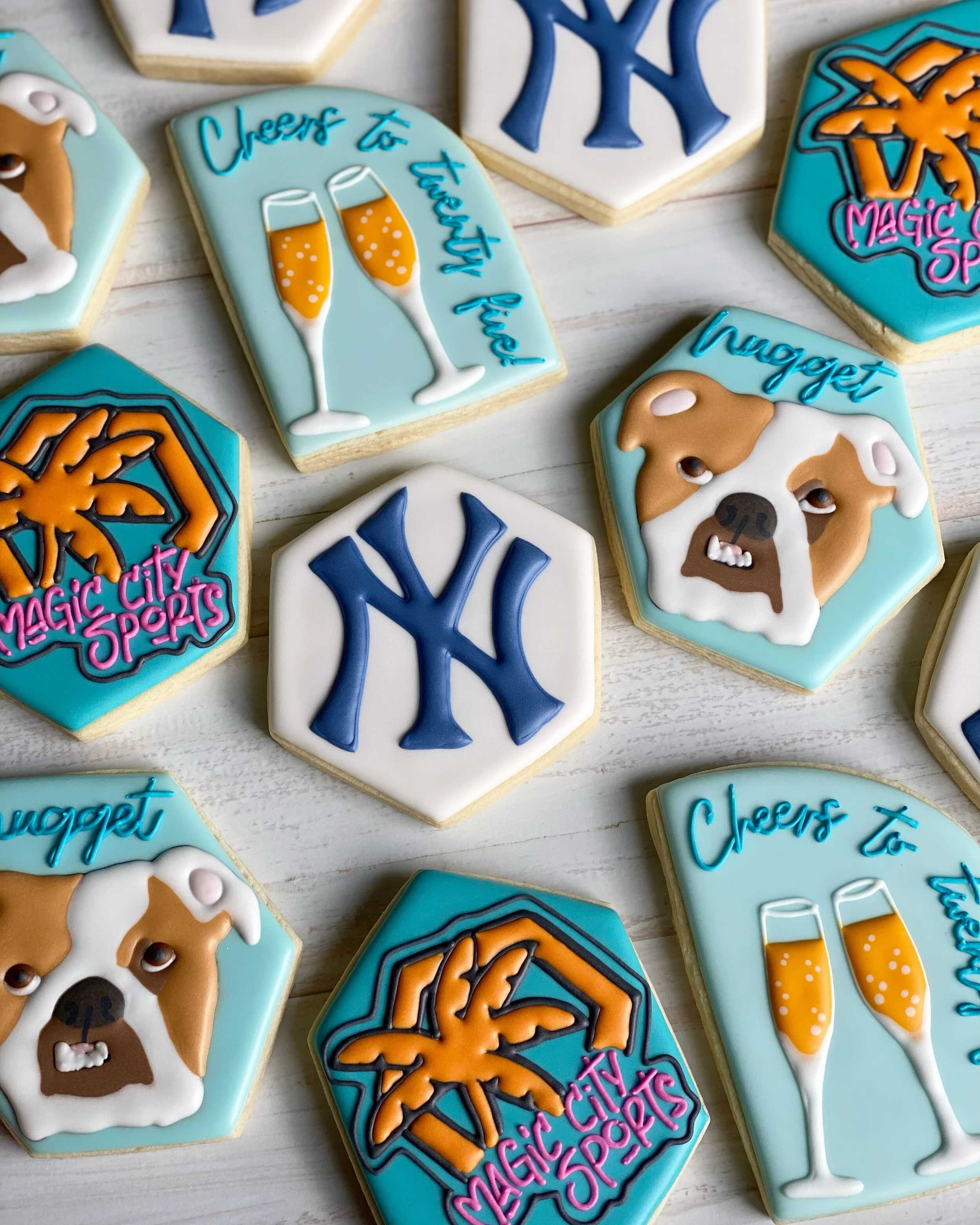 Assorted decorated sugar cookies with New York Yankees and exclusive themed designs, including cheers to tradition, a bulldog face, and city sports logos, laid out on a light wooden surface.