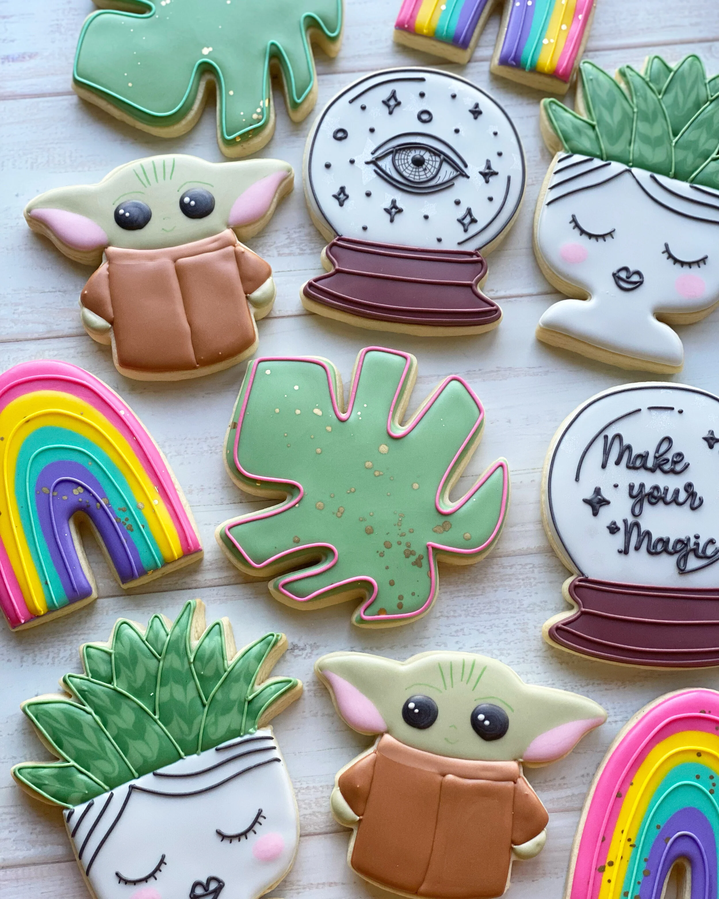Decorated cookies in the shapes of Baby Yoda, a rainbow, a moon with an eye, palm leaves, and a unicorn with closed eyes, on a white wooden surface.