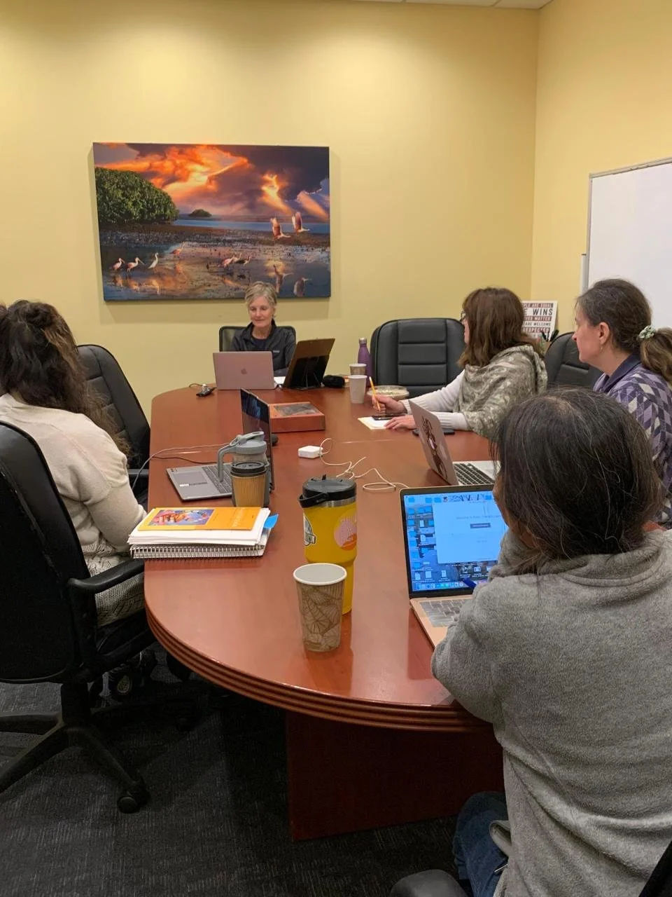 A group of five women sitting around a conference table in a meeting room, engaging in a discussion. The woman at the head of the table is speaking, with a laptop in front of her. There are notebooks, coffee cups, and laptops on the table. A large, vibrant landscape painting hangs on the yellow wall behind her.