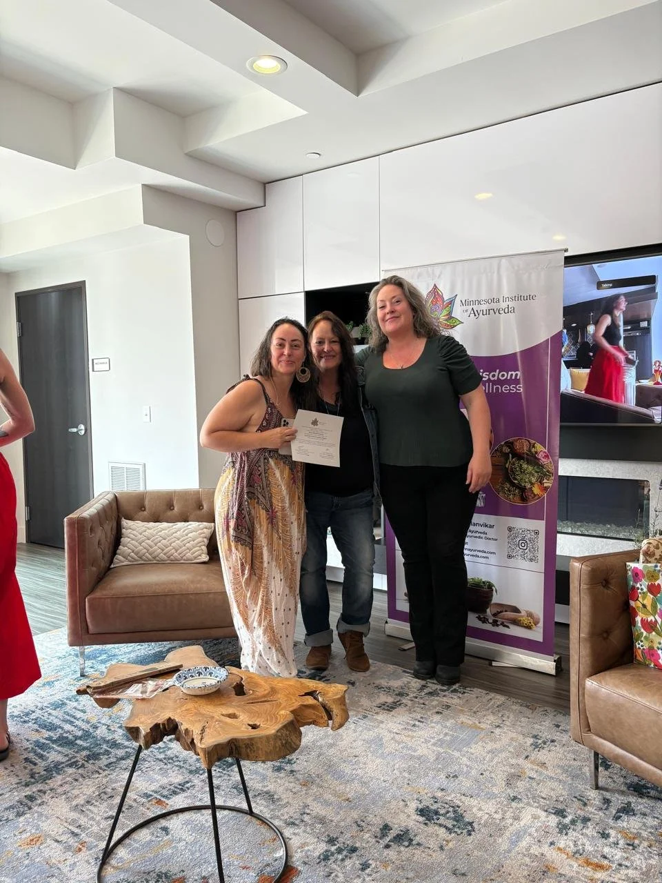 Three women posing together indoors, with one holding a certificate. Behind them is a banner for the Minnesota Institute of Ayurveda. To the left, part of a person in a red dress is visible, and on the right, a floral armchair and small wooden table with decorative items are seen.
