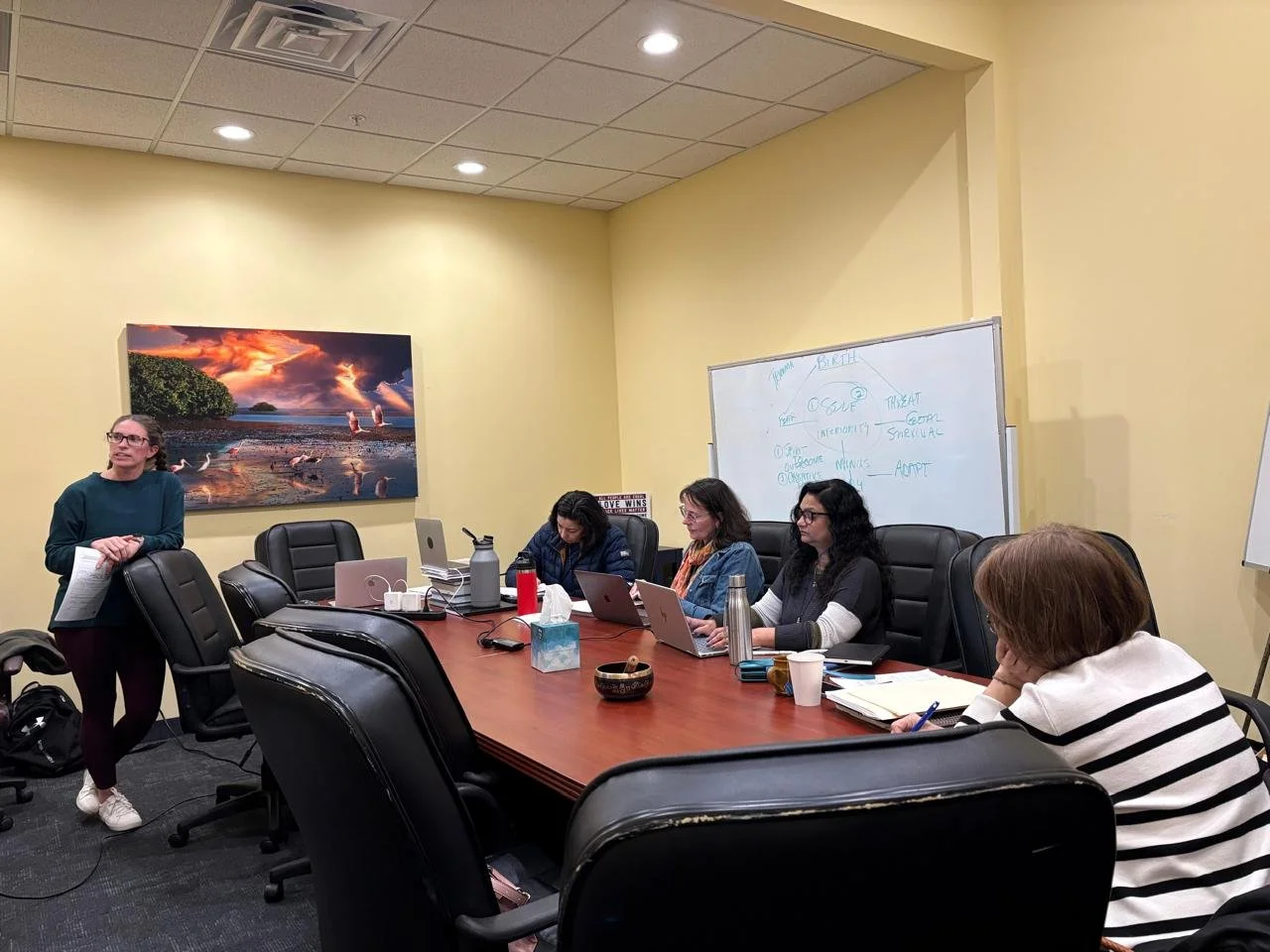 A woman in a green sweater standing and speaking to a group of four women seated at a conference table in a meeting room. The women have laptops and notebooks, and there is a whiteboard with notes behind them. A large framed landscape photograph hangs on the wall.