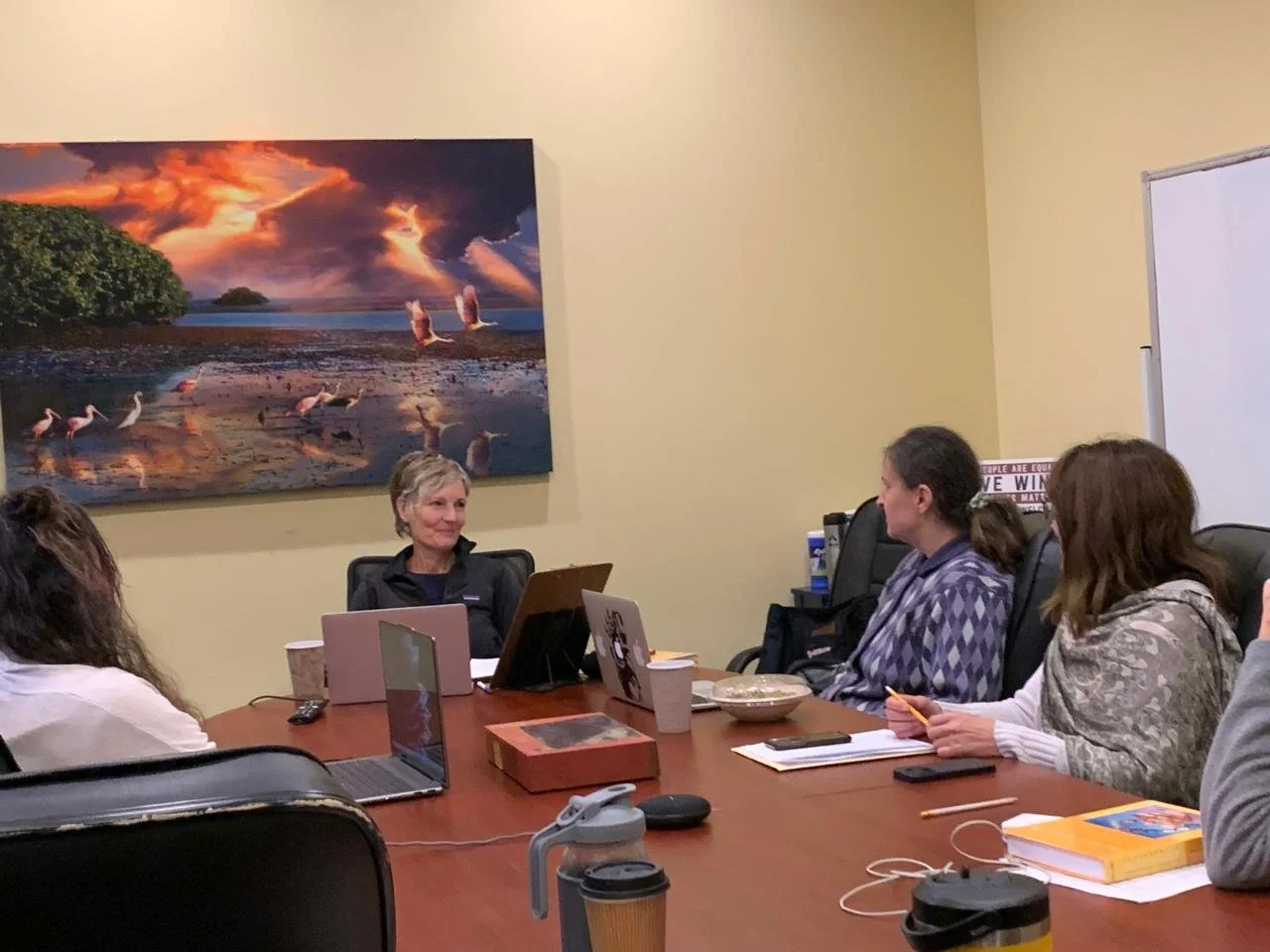 A group of women sitting around a conference table, engaged in a discussion. The woman at the head of the table is smiling. There are laptops, notebooks, and cups on the table. A large landscape painting of a sunset over a body of water with birds is on the wall behind them.