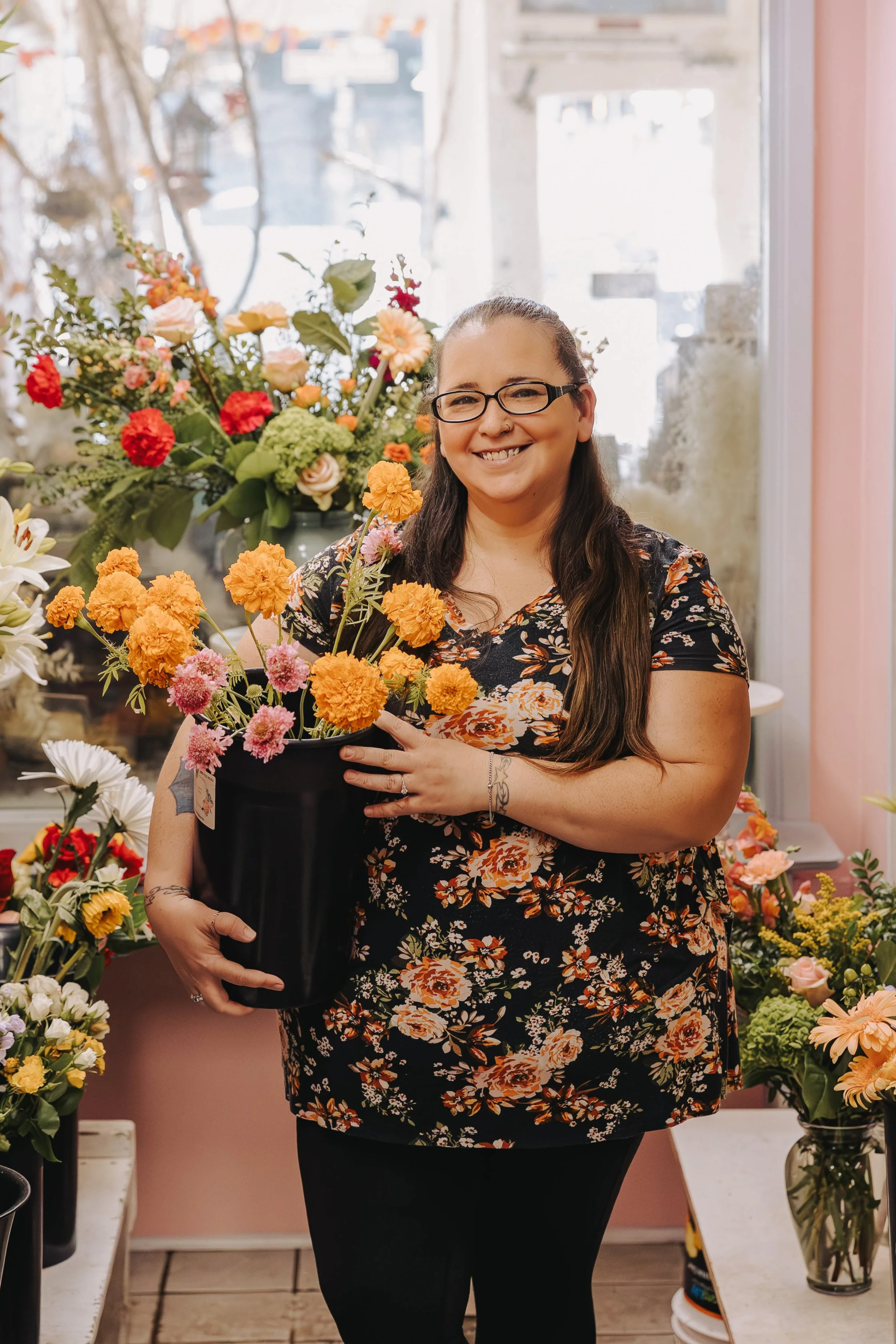 A woman with glasses and long dark hair wearing a striped pink and white shirt, standing in front of a brick wall, smiling at the camera.