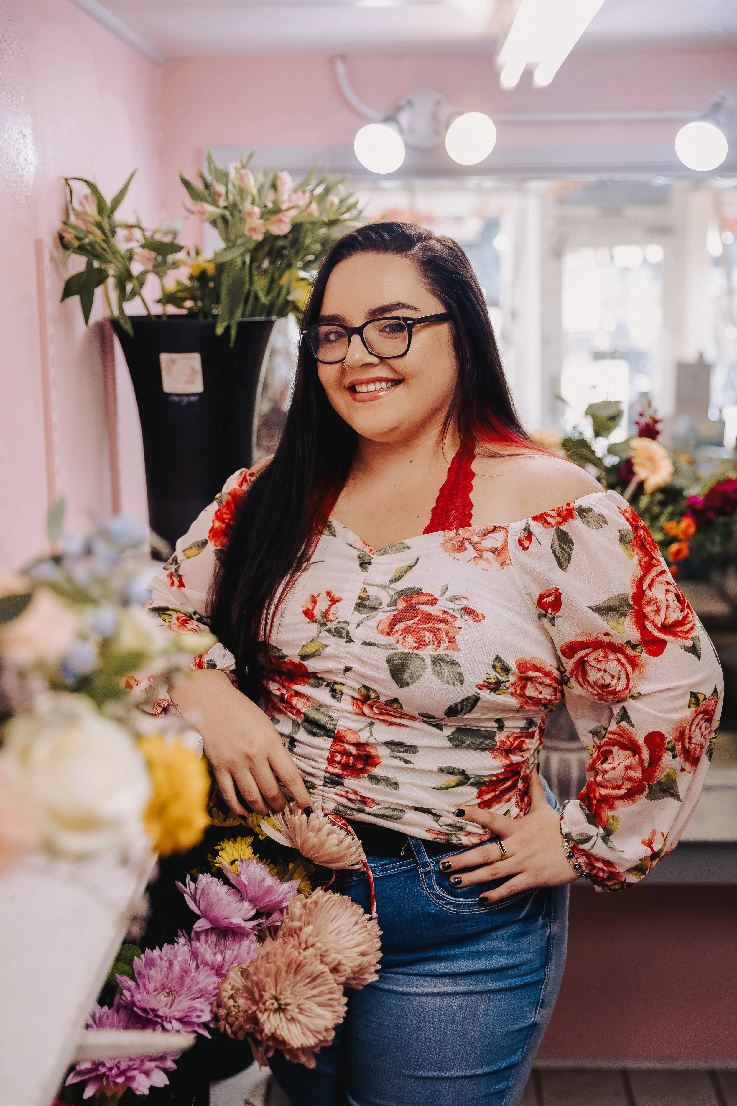 A woman with long wavy brown hair, glasses, and earrings, standing in front of a brick wall, smiling and wearing a teal shirt and dark jeans.