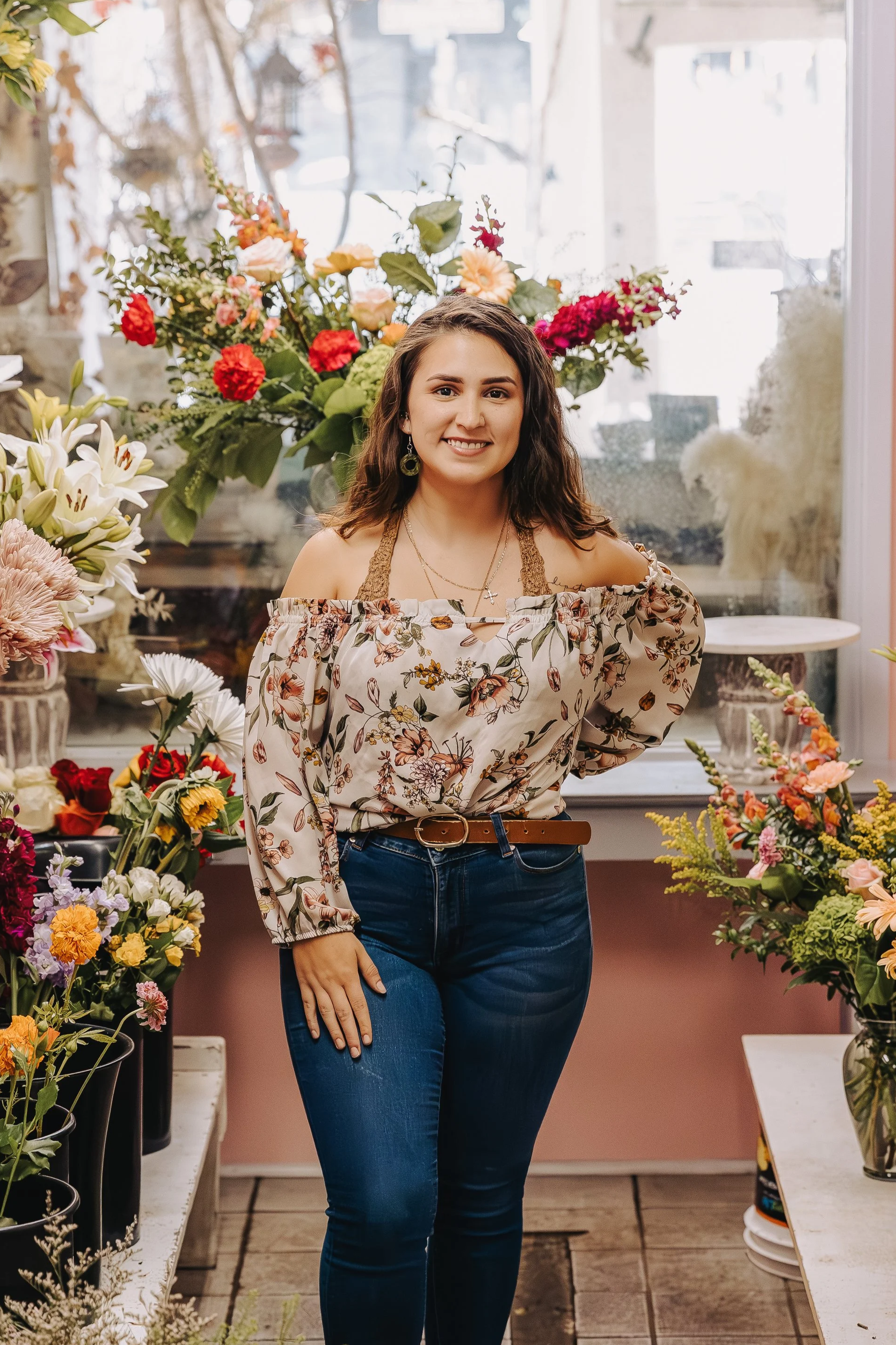 A young woman with long dark wavy hair wearing a tan hat, white blouse, and blue jeans, standing against a brick wall.