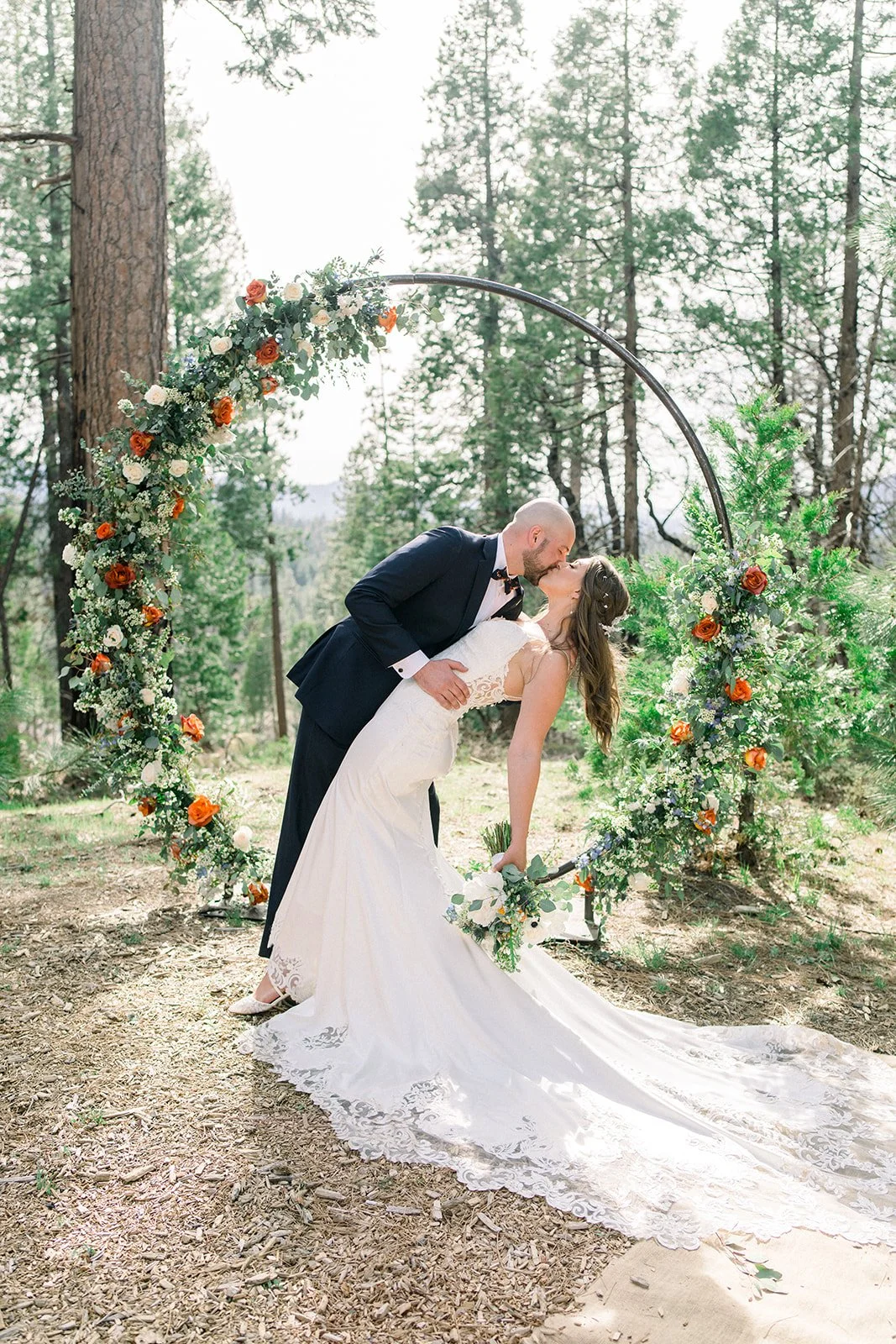Groom dipping bride for kiss in front of a round floral arch made of orange, white, and blue flowers, groveland ca