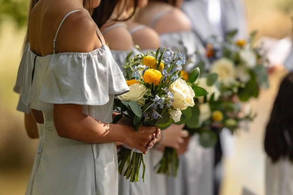 bridesmaid holding a bouquet made of yellow ranunculus, white roses, and blue delphinium, murphys ca wedding florist