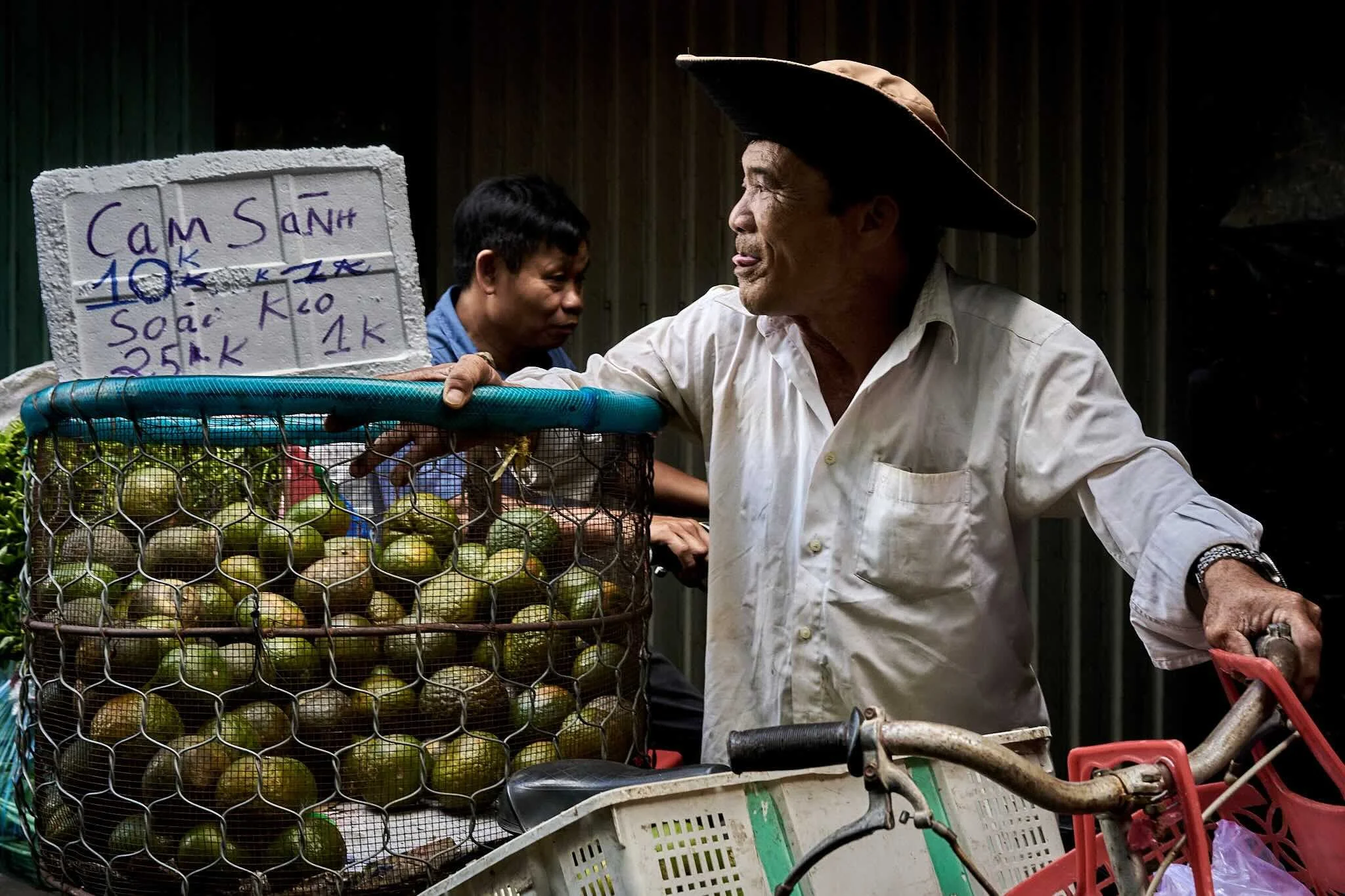 A man wearing a wide-brimmed hat and white shirt interacts with a woman at a fruit stand. The stand has a basket filled with green and yellow fruits, and a sign with handwritten prices in Vietnamese.