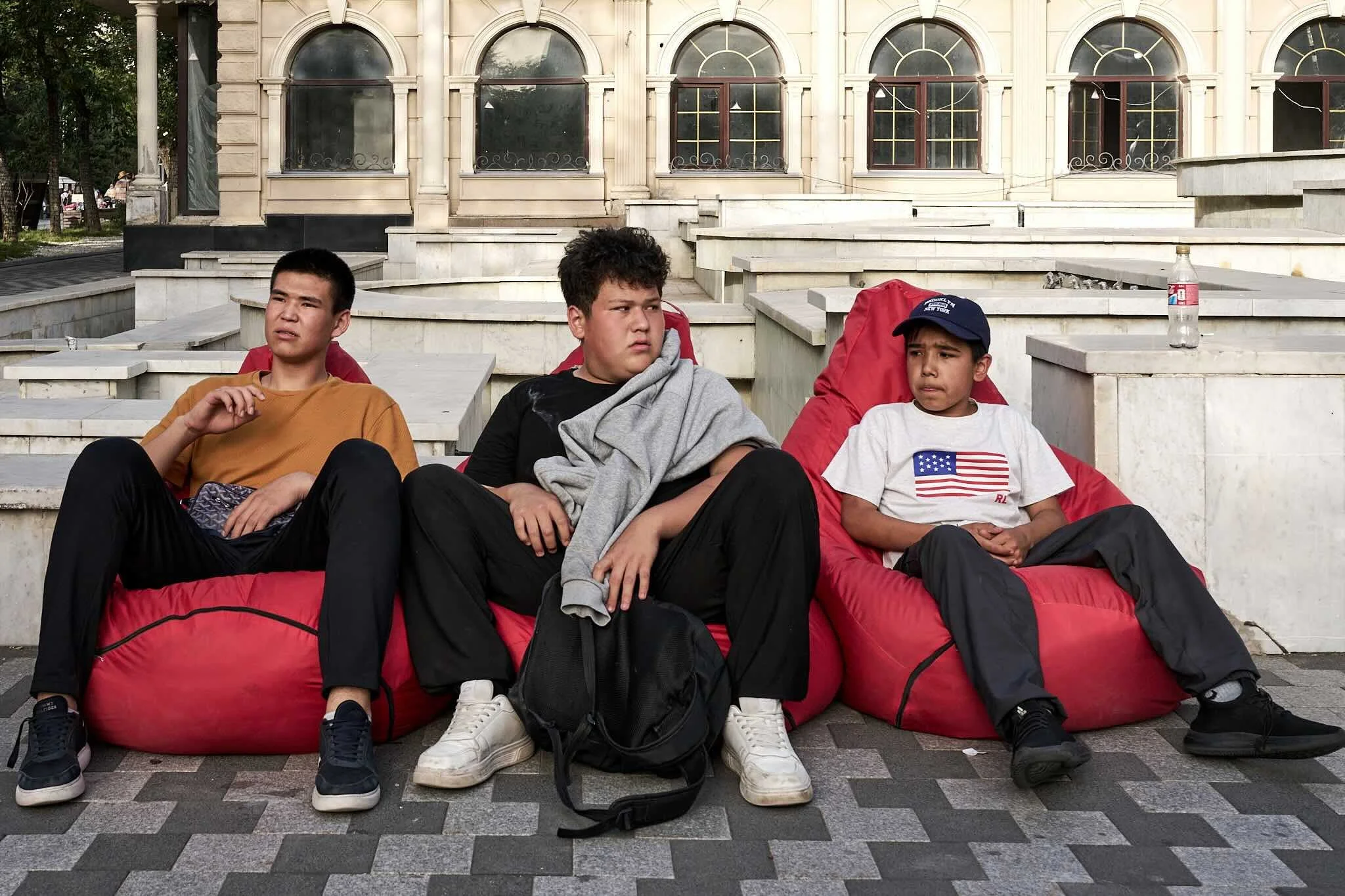 Jerzy Jaworek, Street and Travel Photography, Kazakhstan, Almaty. Three boys sitting on large red bean bag chairs outdoors in front of a building with arched windows and columns.