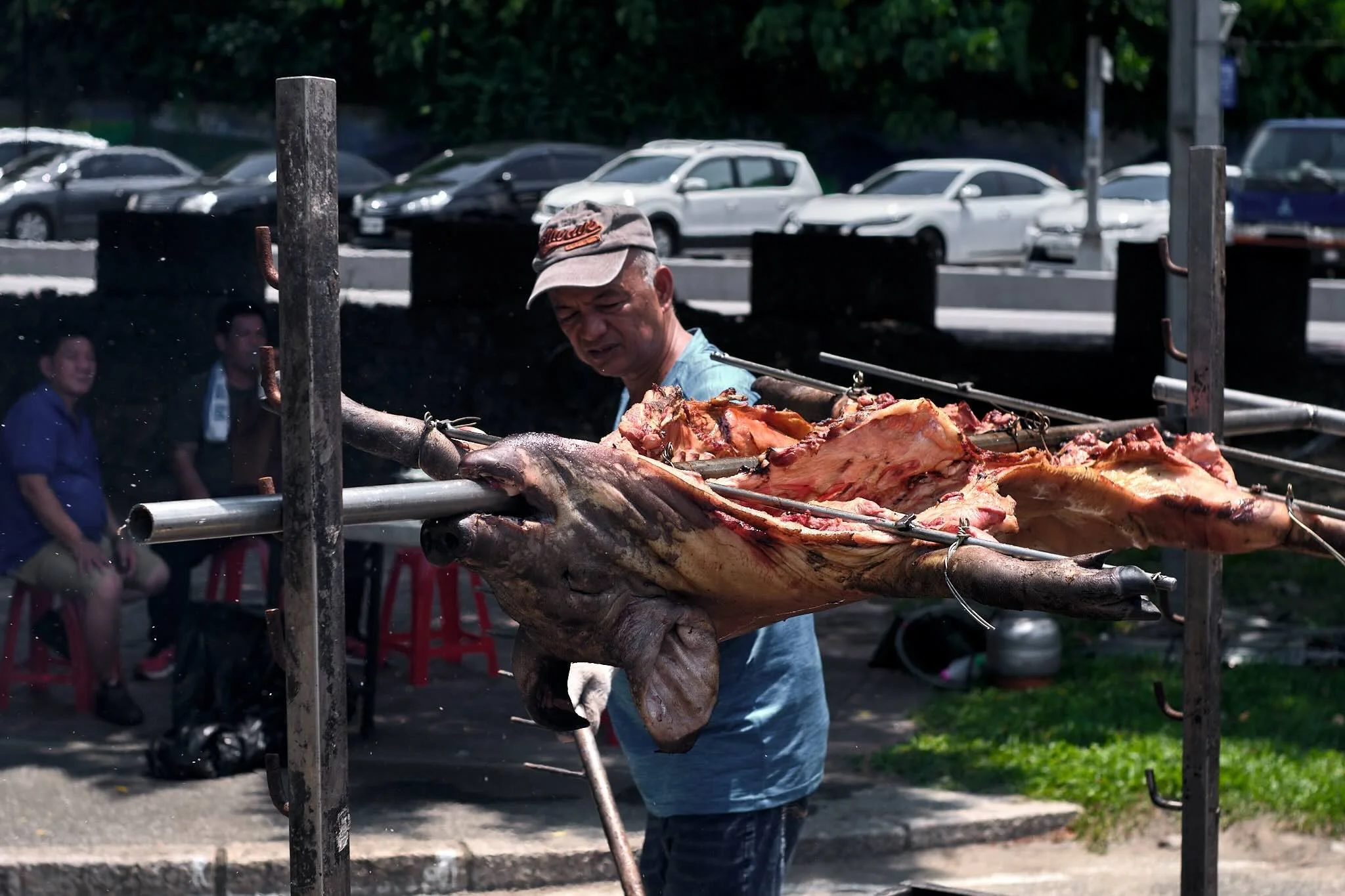 Man wearing a gray cap and blue shirt grilling a large roasted animal, possibly a pig, on a spit outdoors with parked cars and people in the background.