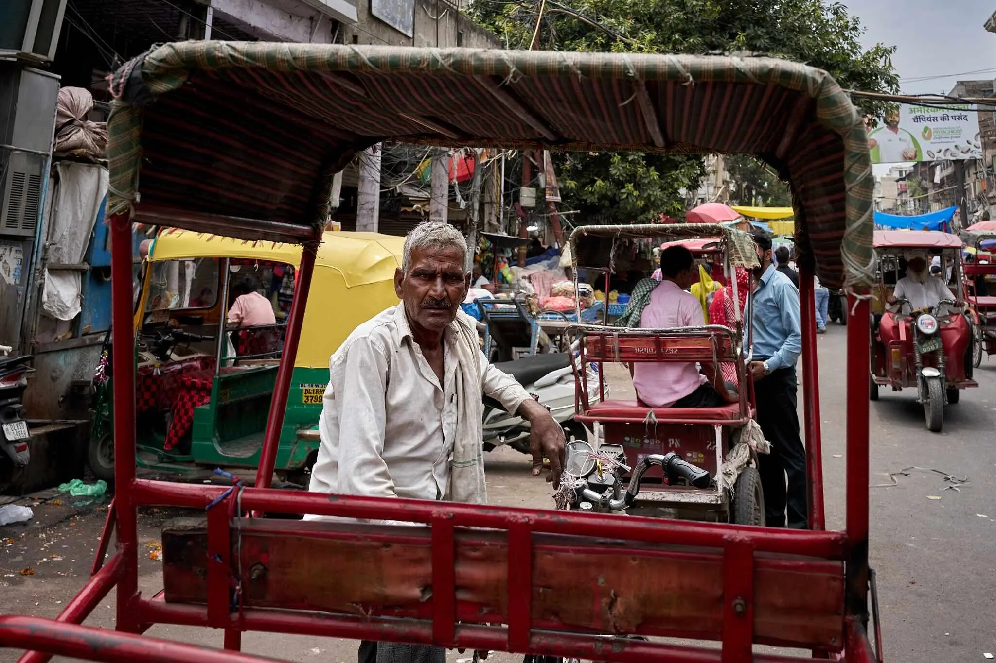 Jerzy Jaworek, Street and Travel Photography, India, Delhi. A busy street scene in India with pedestrians, auto rickshaws, and a man sitting on a red vehicle in the foreground. Market stalls and colorful umbrellas are visible in the background.